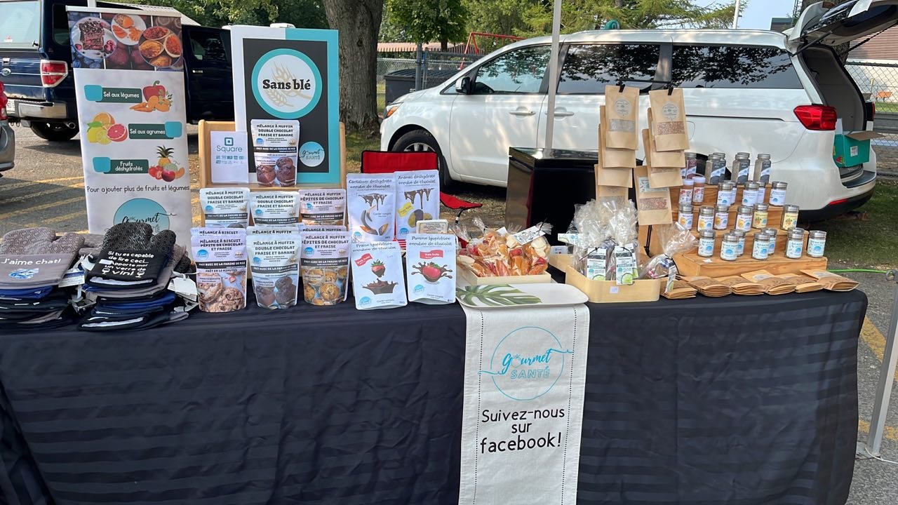 Un stand de produits alimentaires et de merchandising, avec des sachets de thé, des pots de miel, des brochures, des sacs en papier, des vêtements et une bannière promotionnelle, situé dans un parking en extérieur.