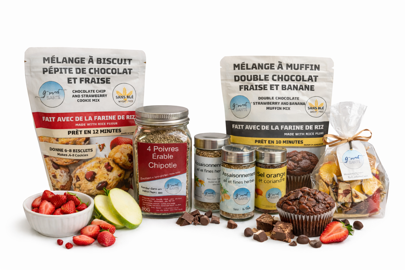 Différents ingrédients et préparations pour pâtisserie, incluant des sachets de mélanges à biscuits et muffins, des épices en petits pots, un bol de fraises, des tranches de pomme, des morceaux de chocolat, et une petite poche de bonbons, sur un fond blanc.