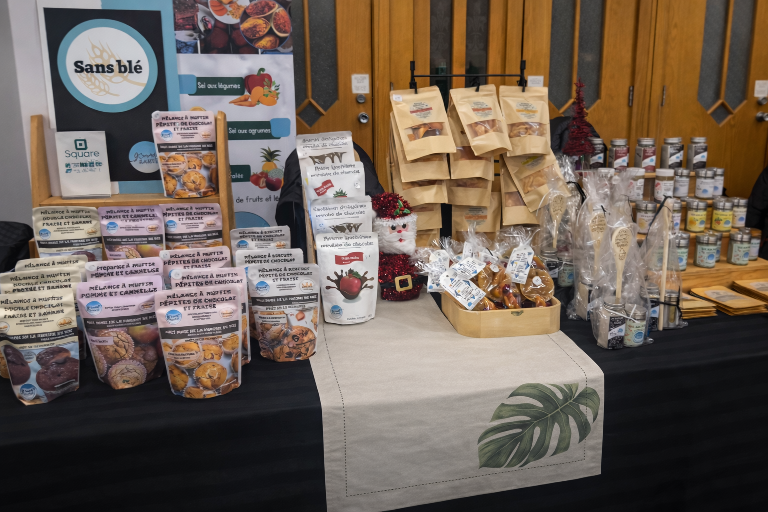 Un stand de produits alimentaires sans gluten avec des paquets de biscuits, des produits en bouteille, et des sachets en papier. Décoration festive avec un petit bonhomme de neige. Mises en page organisées avec panneaux et étiquettes.