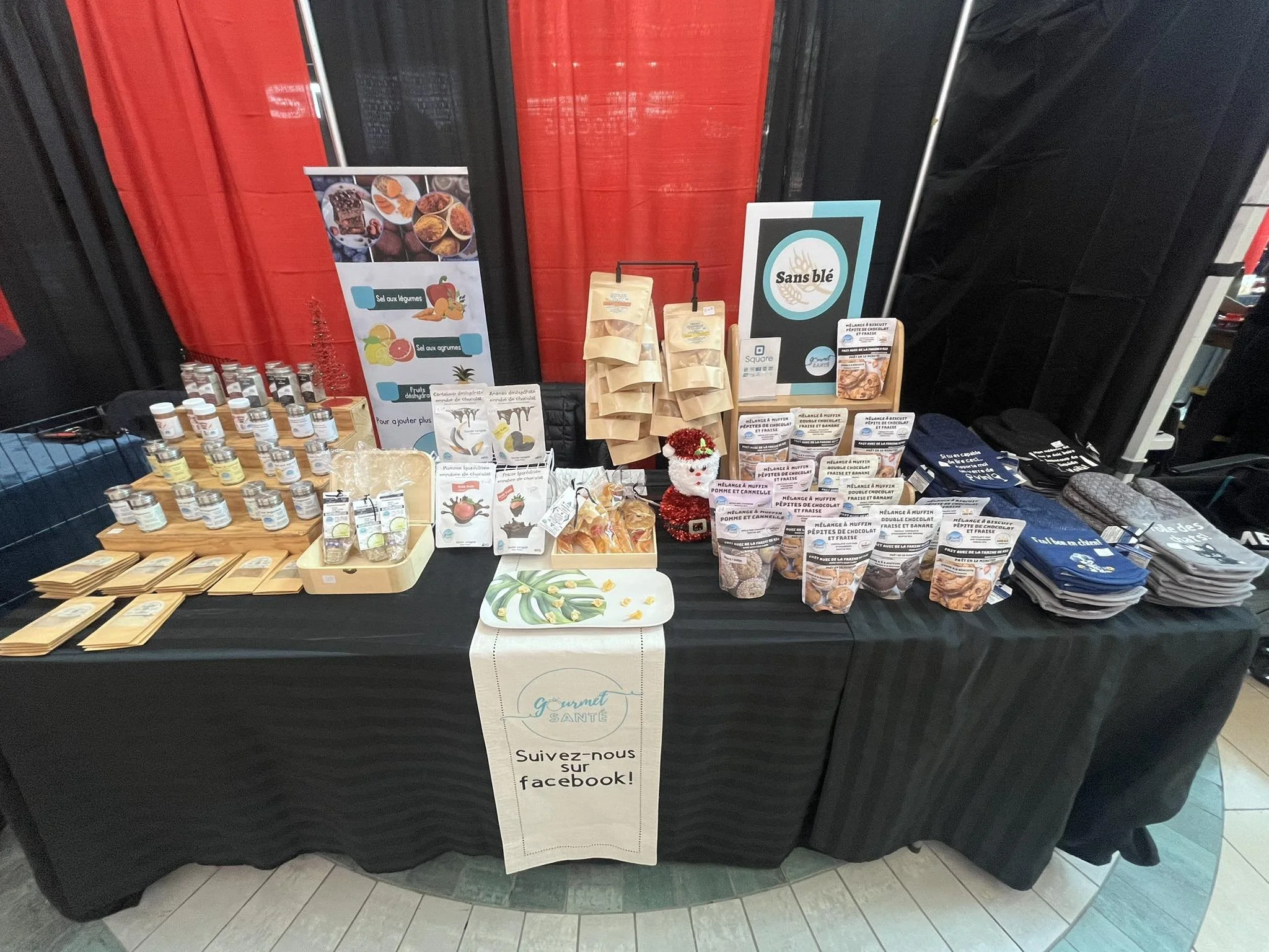 Table de présentation de produits alimentaires bio avec emballages, échantillons et brochures, décorée avec un petit bonhomme de Noël, dans un stand de marché ou exposition.