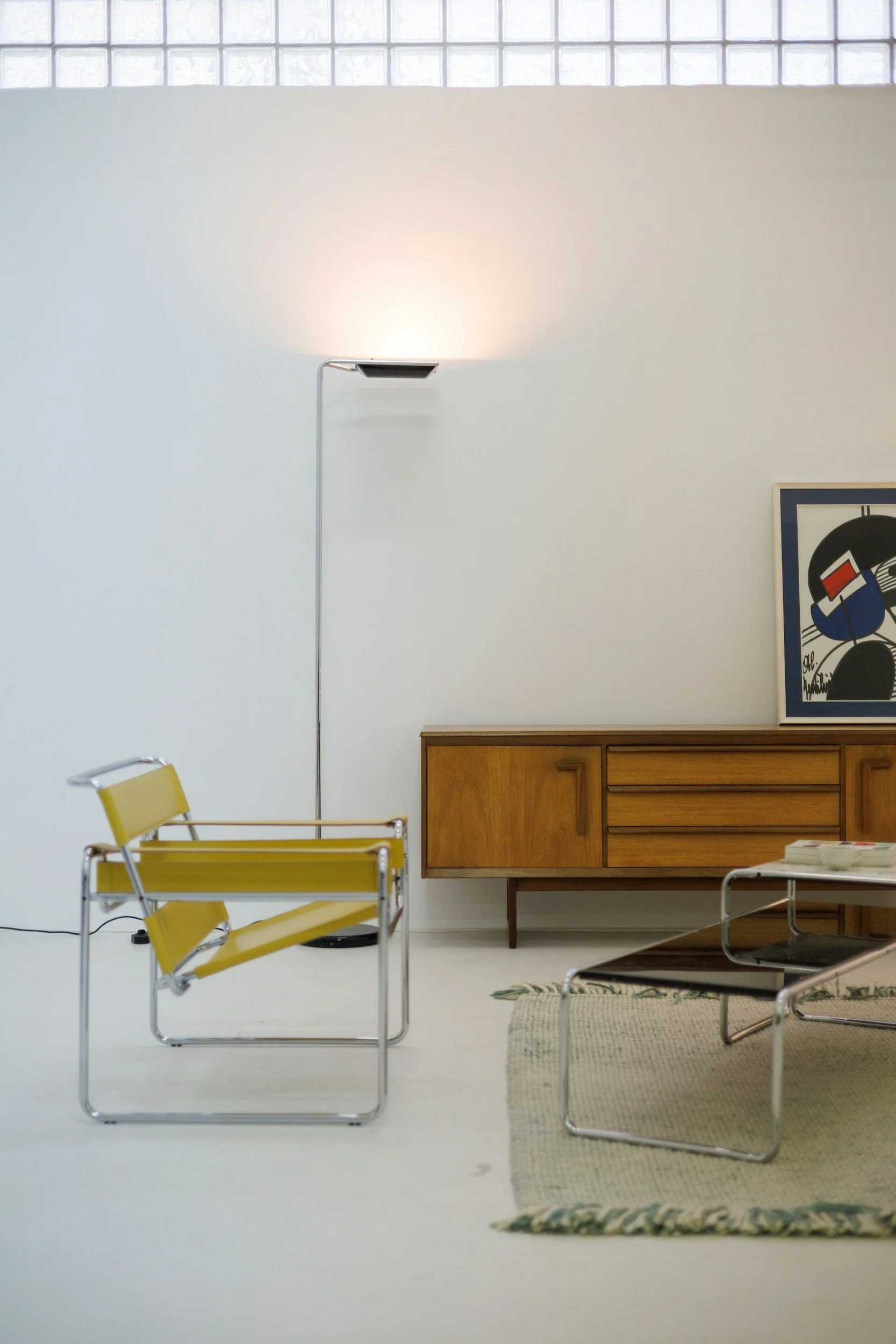 A mid-century modern living room with a yellow lounge chair, a wooden sideboard, a framed abstract artwork, a black coffee table, a floor lamp, and a beige rug.