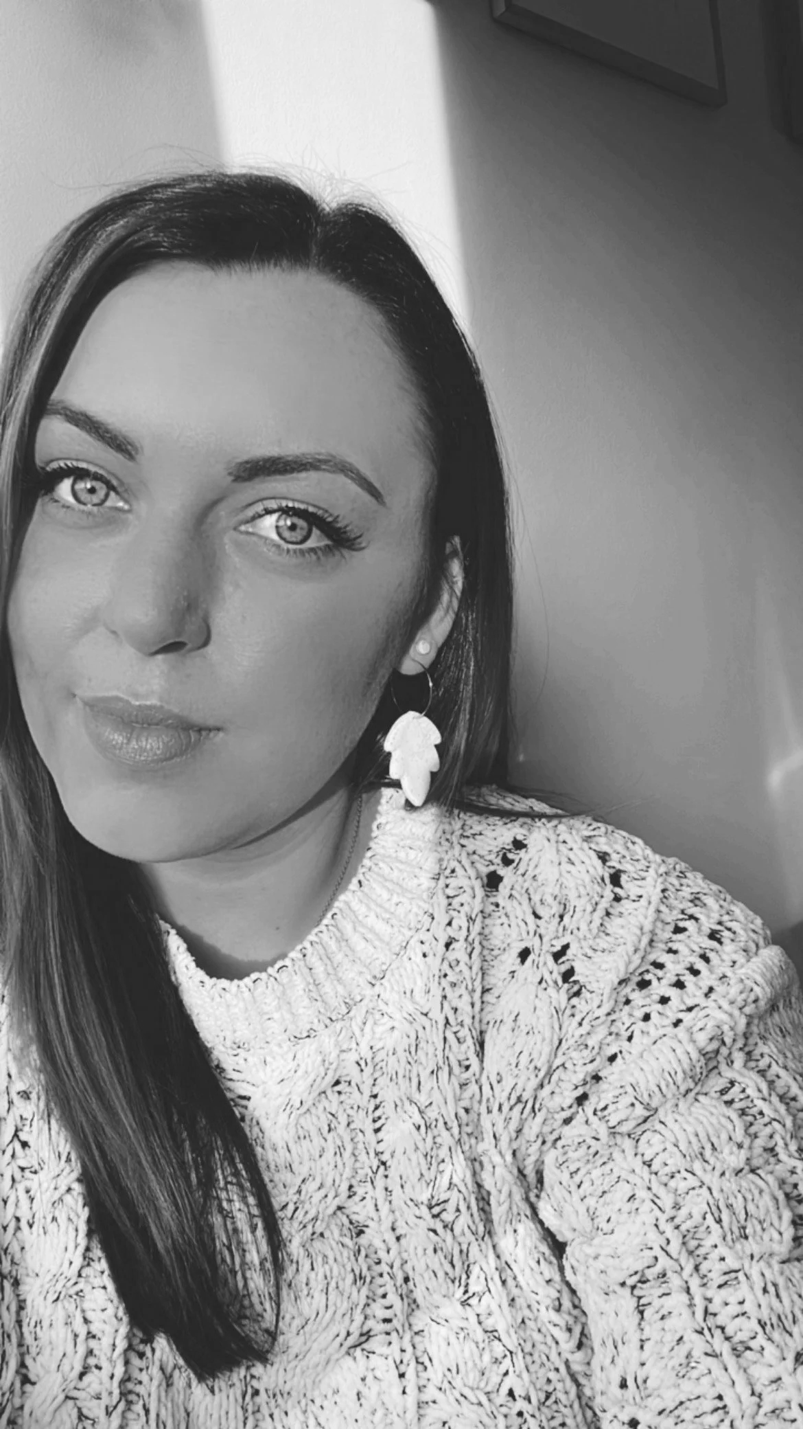 Close-up black and white photo of a woman with light-colored eyes, long dark hair, wearing a patterned sweater and leaf-shaped earrings, taking a selfie indoors with a plain wall and framed picture in background.