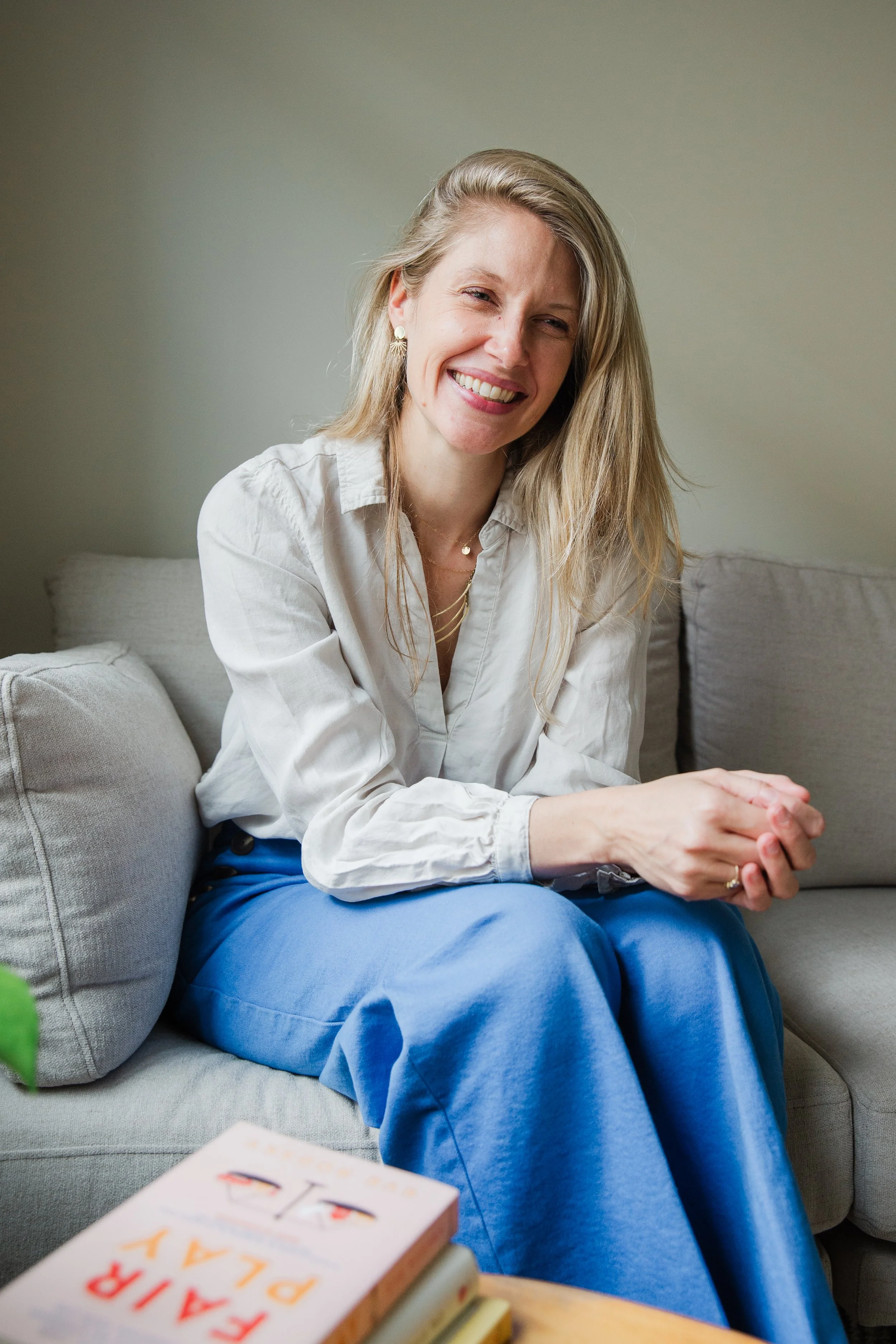 A smiling woman with long blond hair, wearing a white blouse and blue pants, sitting on a light-colored couch in a cozy, softly lit room.