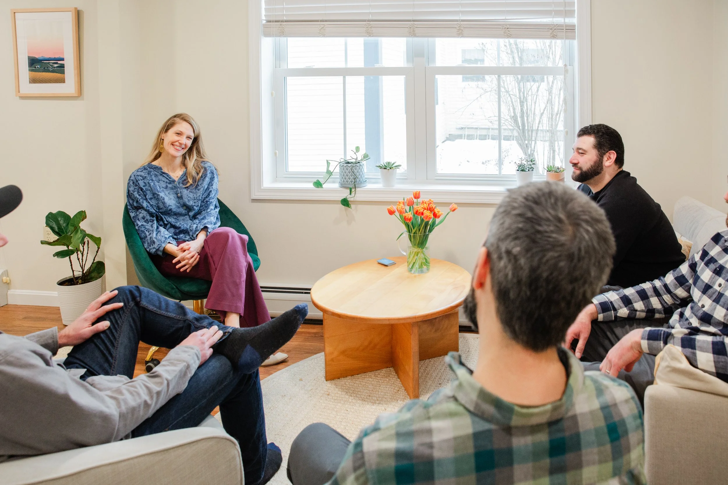 A woman smiling and sitting on a green chair in a living room, surrounded by five people. There is a window behind her with several potted plants on the windowsill and a vase of flowers on the wooden coffee table in front of her. The other people are sitting on sofas and chairs, listening attentively.