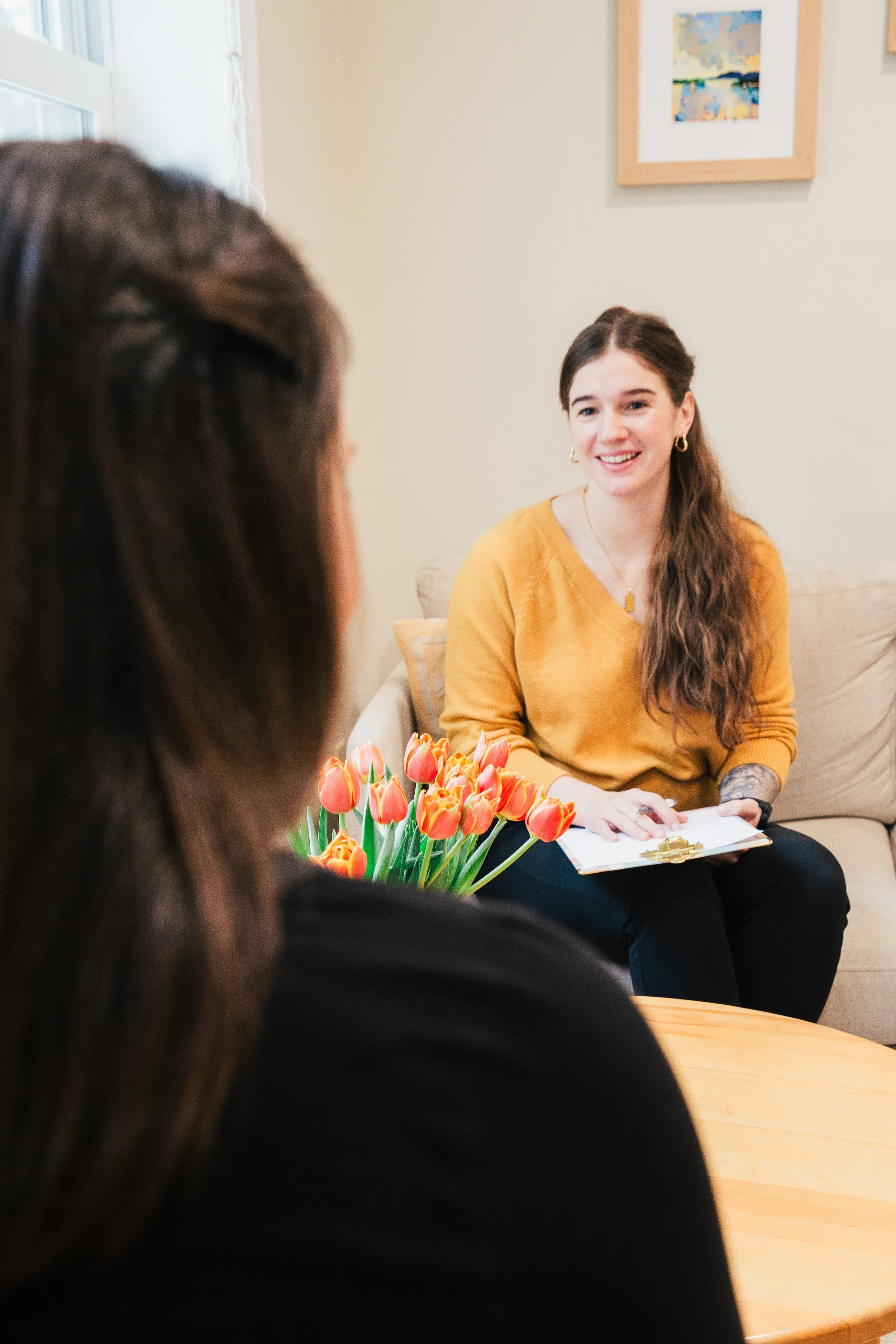 A woman in a yellow sweater sitting on a beige couch, smiling and holding a clipboard, with another woman in the foreground, and a bouquet of orange tulips on a wooden table.