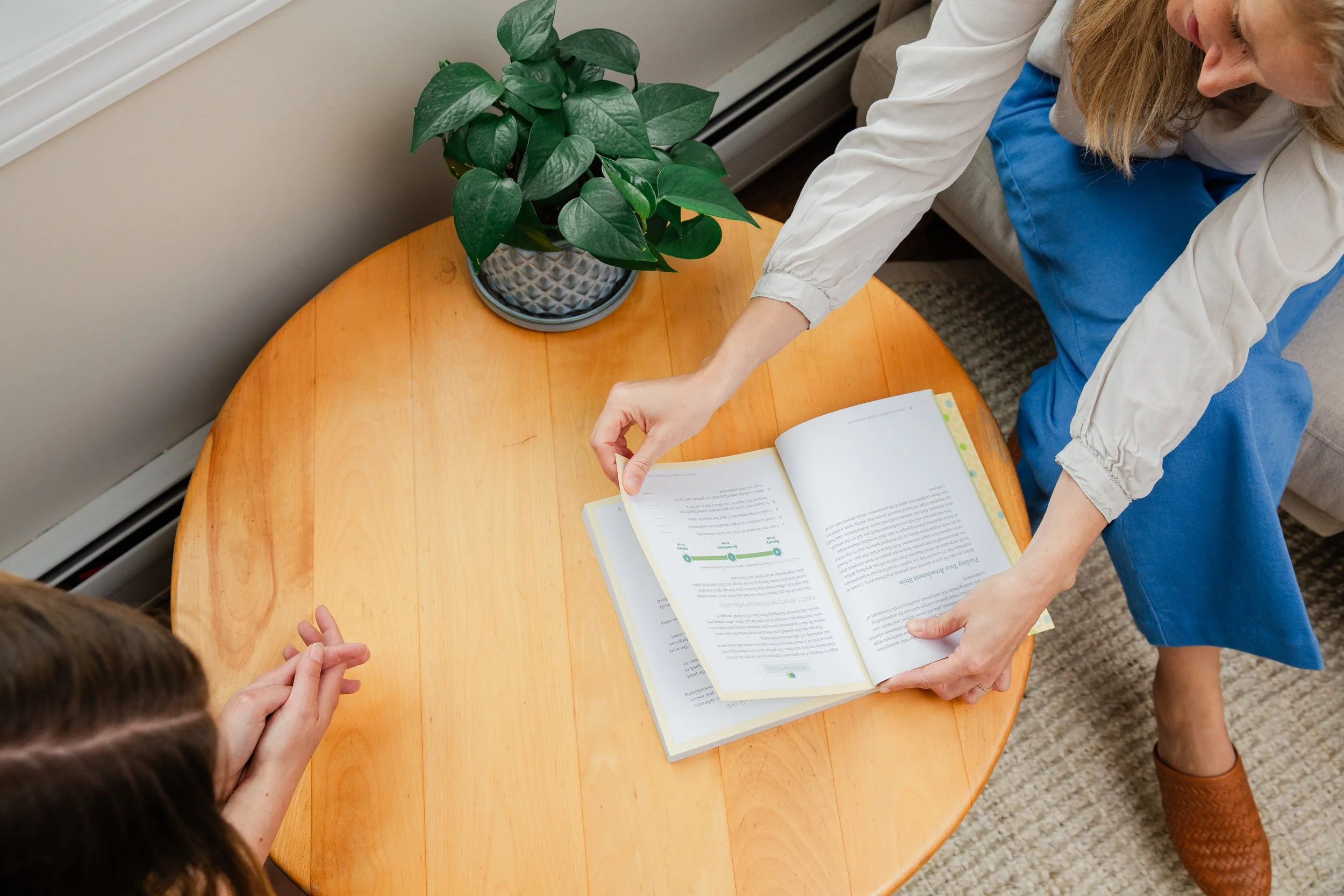 A woman sitting on a sofa shows a book to a child while reaching for the open book on a round wooden table. The table has a potted plant with green leaves on it.