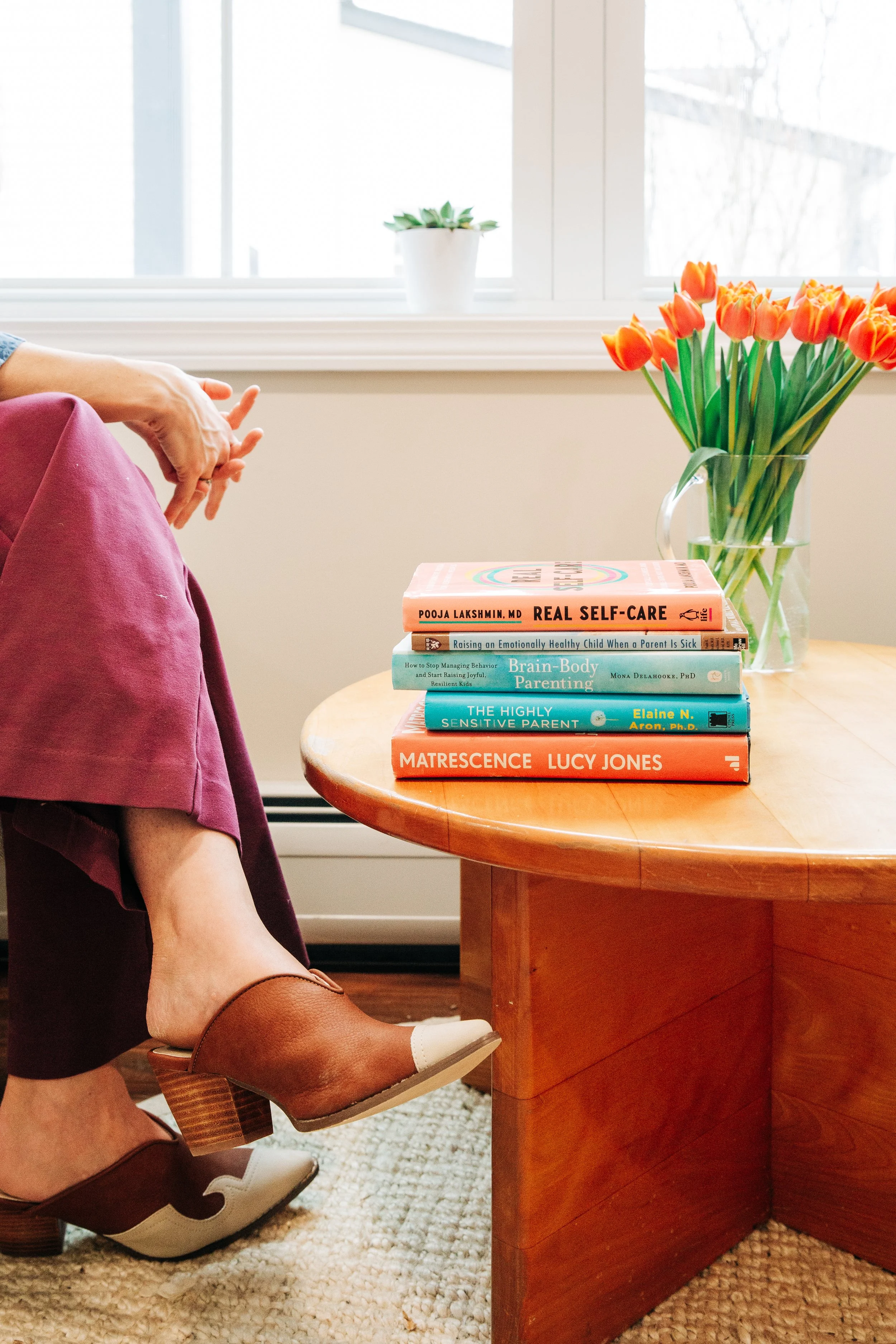 A wooden table with a stack of five books on it, next to a large glass vase filled with orange tulips. In the background, there is a white window with a small potted plant on the windowsill. A person is sitting nearby, wearing maroon pants and brown shoes, with legs crossed.