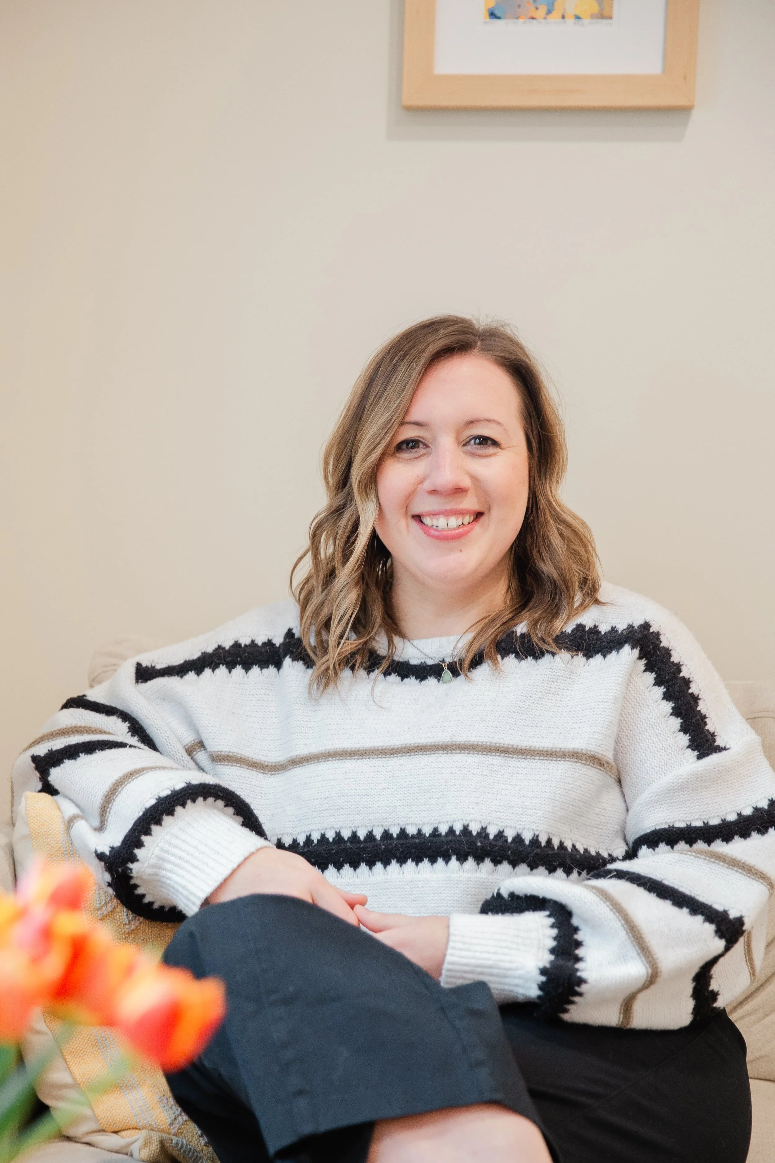 A woman sitting on a beige couch, smiling at the camera, wearing a cream-colored sweater with black and brown stripes, and dark pants, with a framed picture hanging on a light-colored wall behind her.