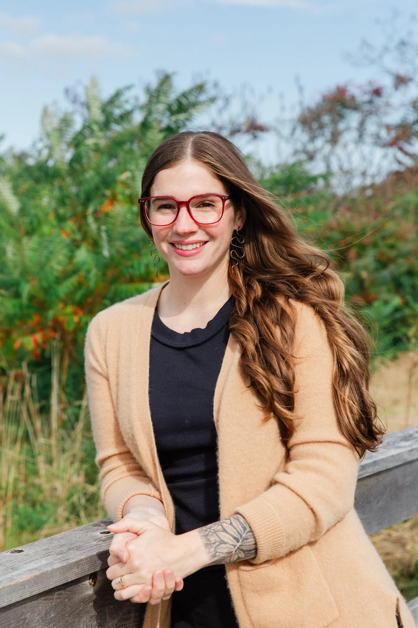 Smiling woman with long, wavy brown hair wearing red glasses, a black top, and beige cardigan standing outdoors with green trees and blue sky in the background.