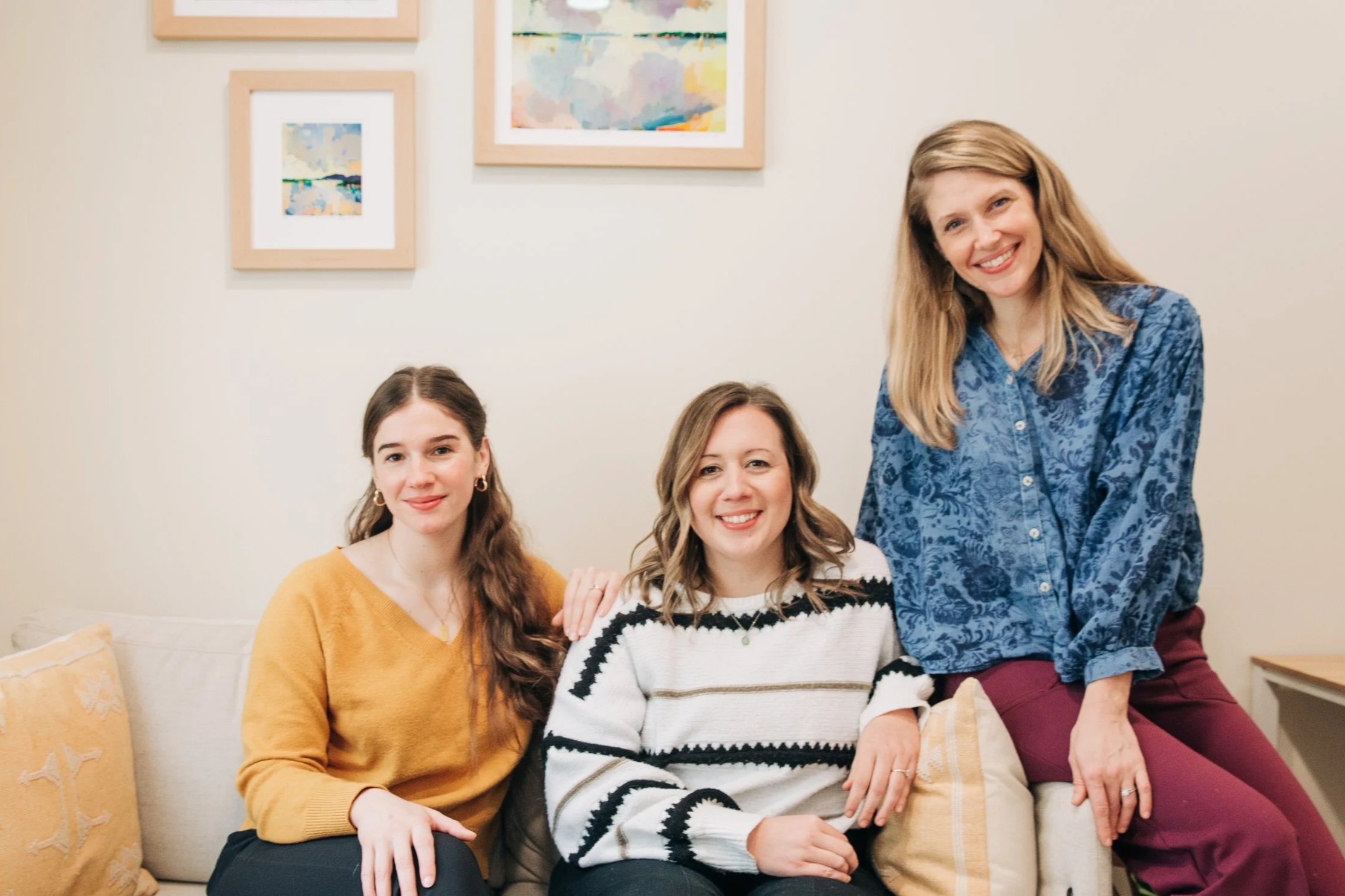 Three women sitting on a beige sofa in a living room, smiling at the camera. The woman on the left wears a mustard-colored sweater, the woman in the middle wears a white and black striped sweater, and the woman on the right wears a blue patterned blouse and maroon pants. There are yellow cushions on the sofa, and framed abstract art paintings hang on the wall behind them.