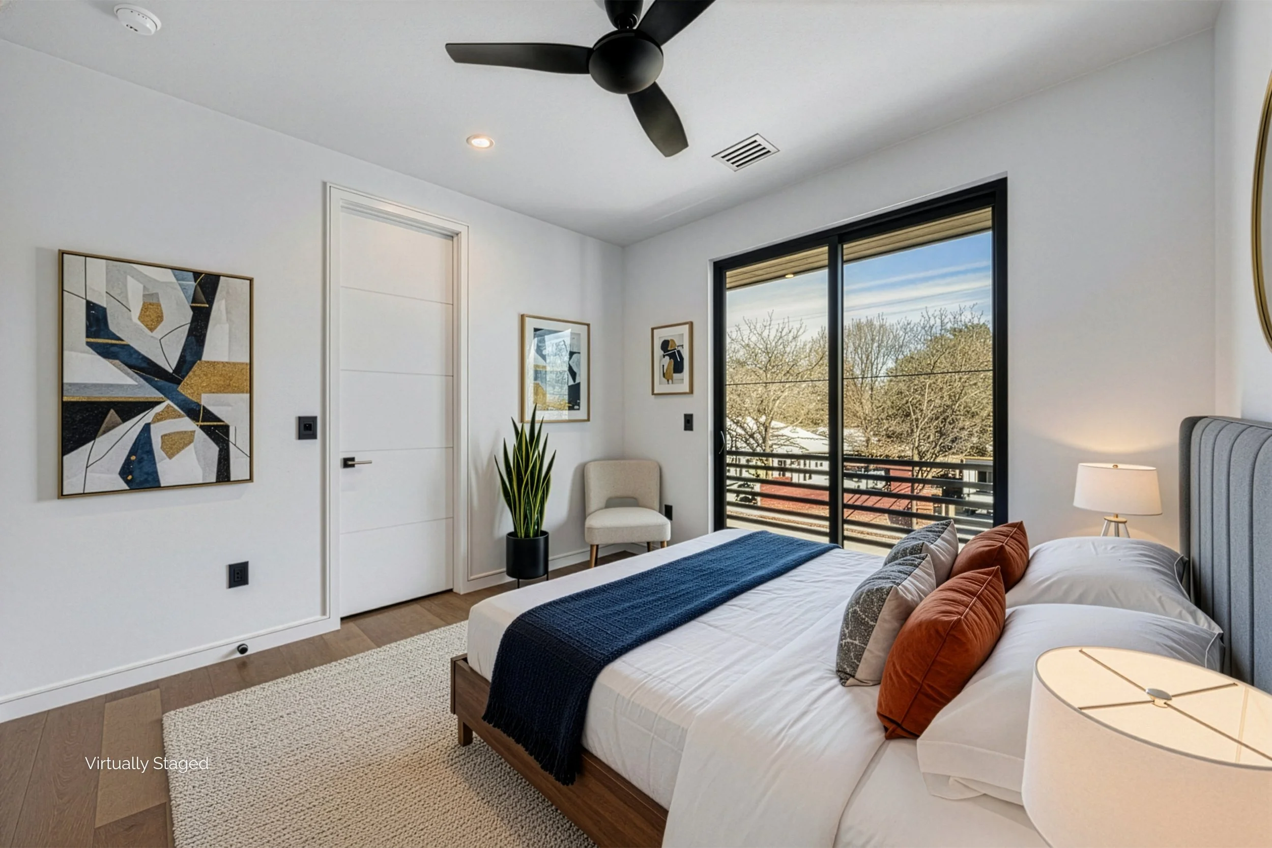 A modern bedroom with a large bed covered in white sheets, multiple pillows, a navy throw, and a wooden bed frame. There's a nightstand with a lamp on the right, a sliding glass door leading to a balcony, and artwork on the walls. A ceiling fan and a