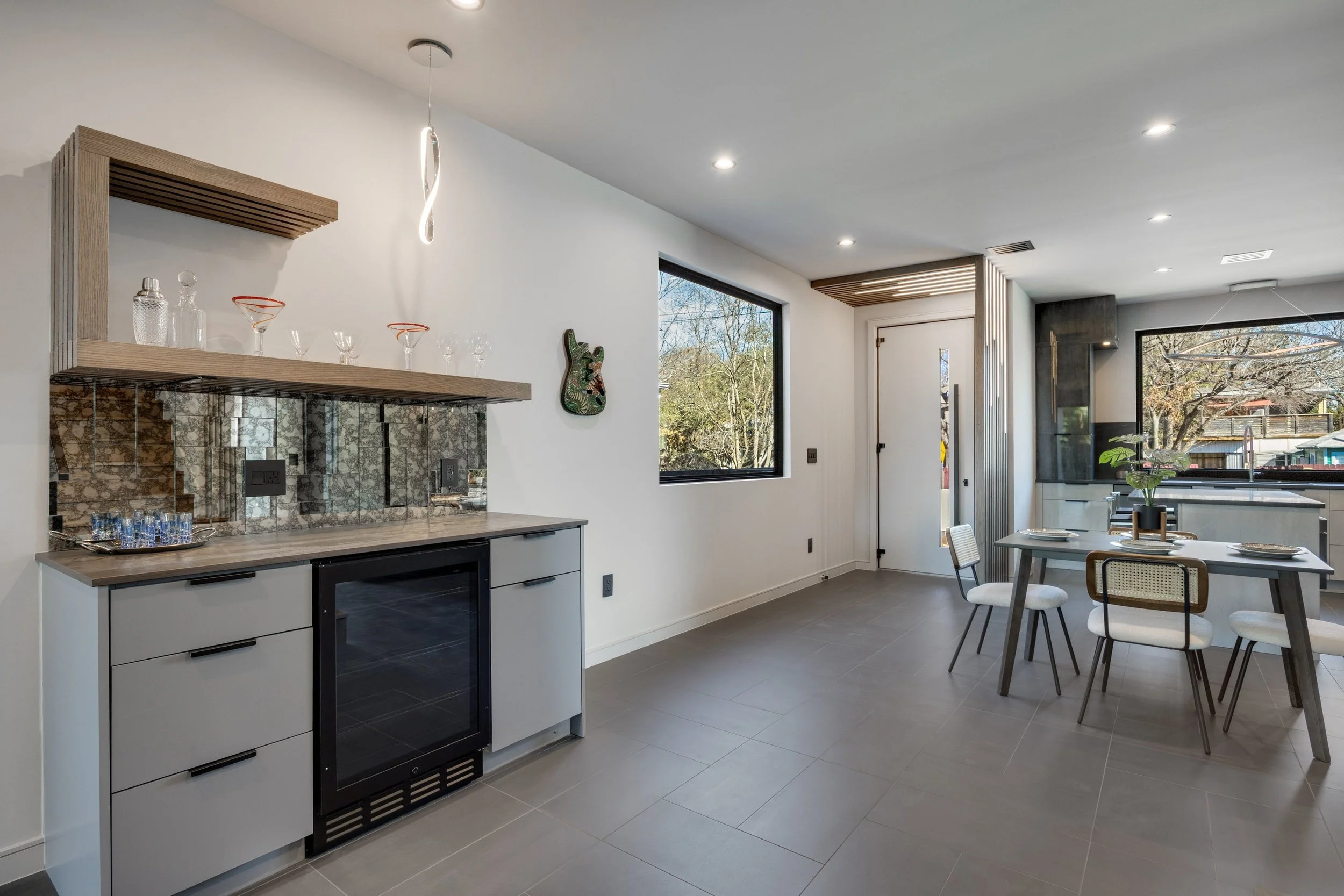 Modern dining area with a gray table, white chairs, and a potted plant, large windows showing trees outside, and a kitchenette with a black and gray backsplash.