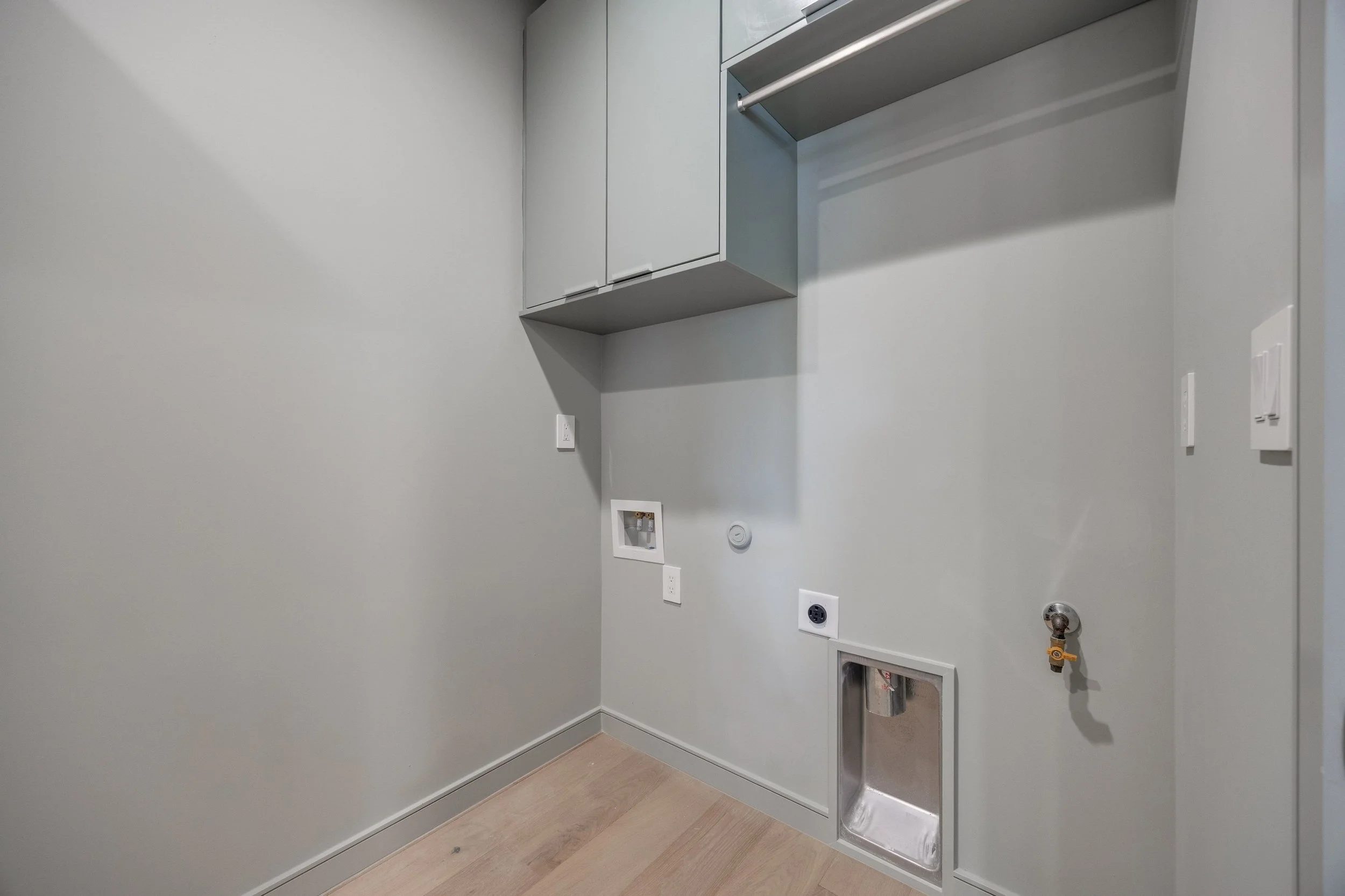 Empty laundry room with light gray walls, a small metallic laundry sink, an open laundry hookup, and a closed gray cabinet above.