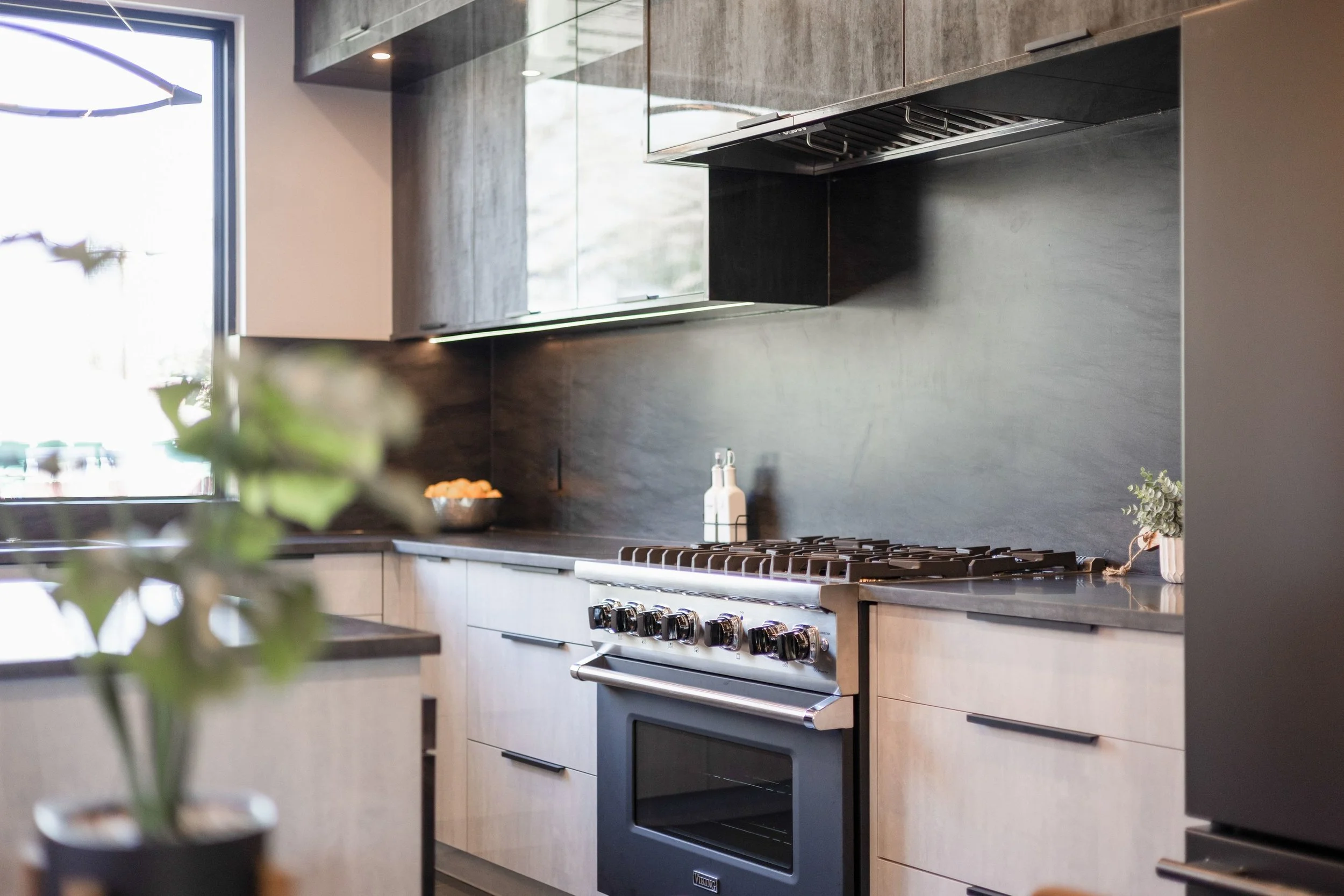 Modern kitchen with black and light wood cabinets, a stainless steel stove, and a dark backsplash. A window with natural light and some plants and bottles on the countertops.