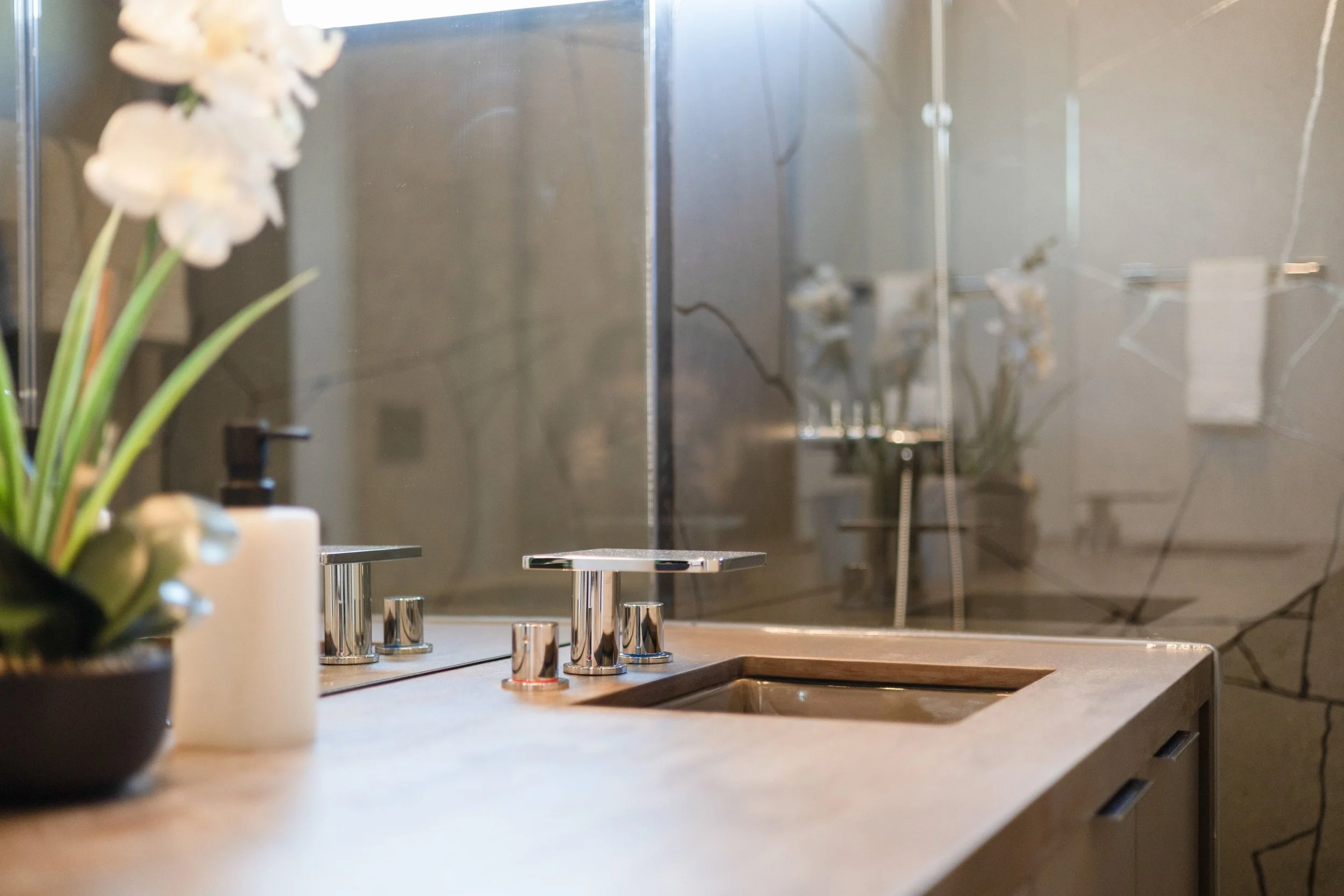 A modern bathroom countertop with a built-in sink, a soap dispenser, a flower pot with white orchids, and a large mirror reflecting the interior.