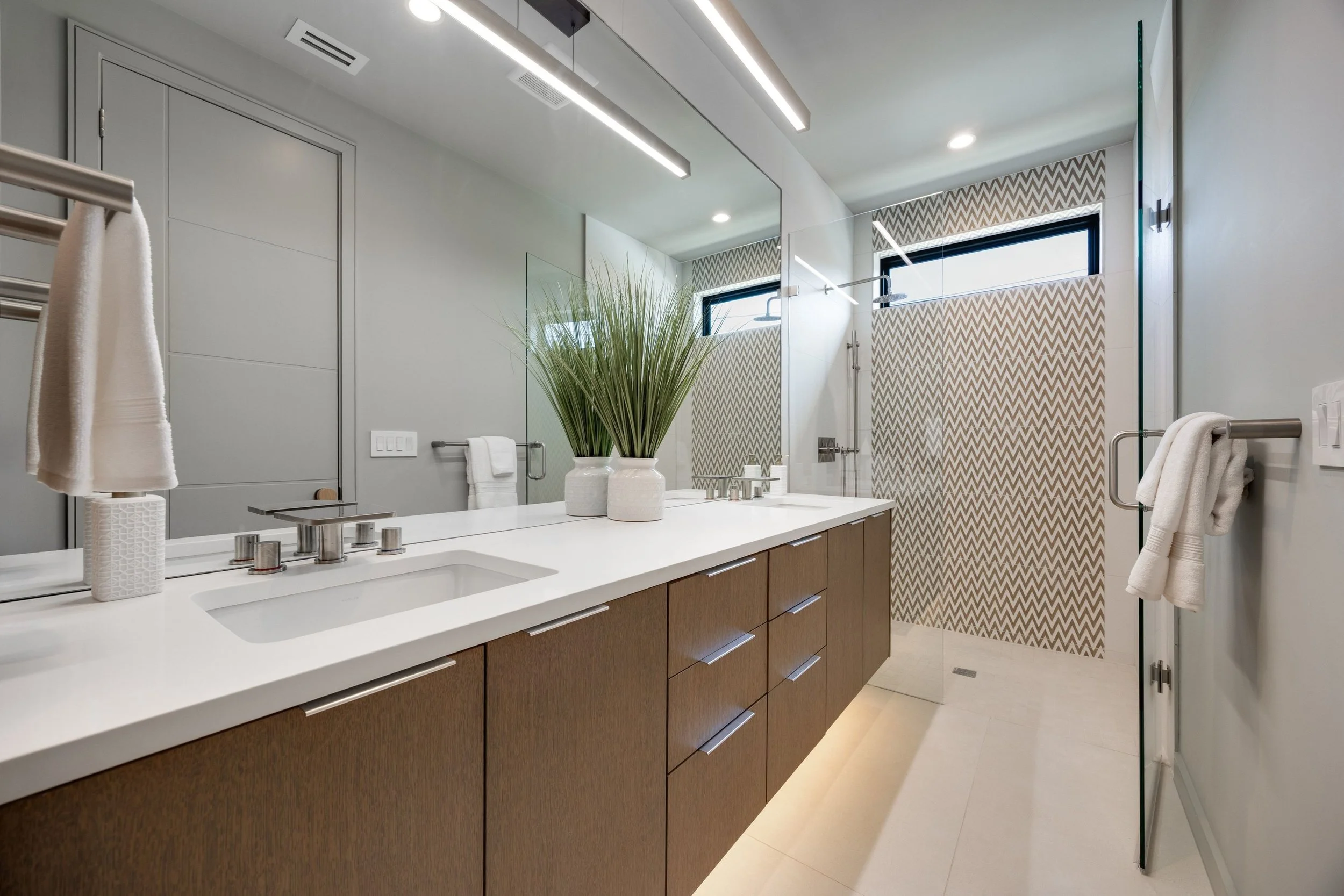 Modern bathroom with a large mirror, wooden vanity with white countertop, potted plants, towel racks, a patterned shower area, and horizontal windows.