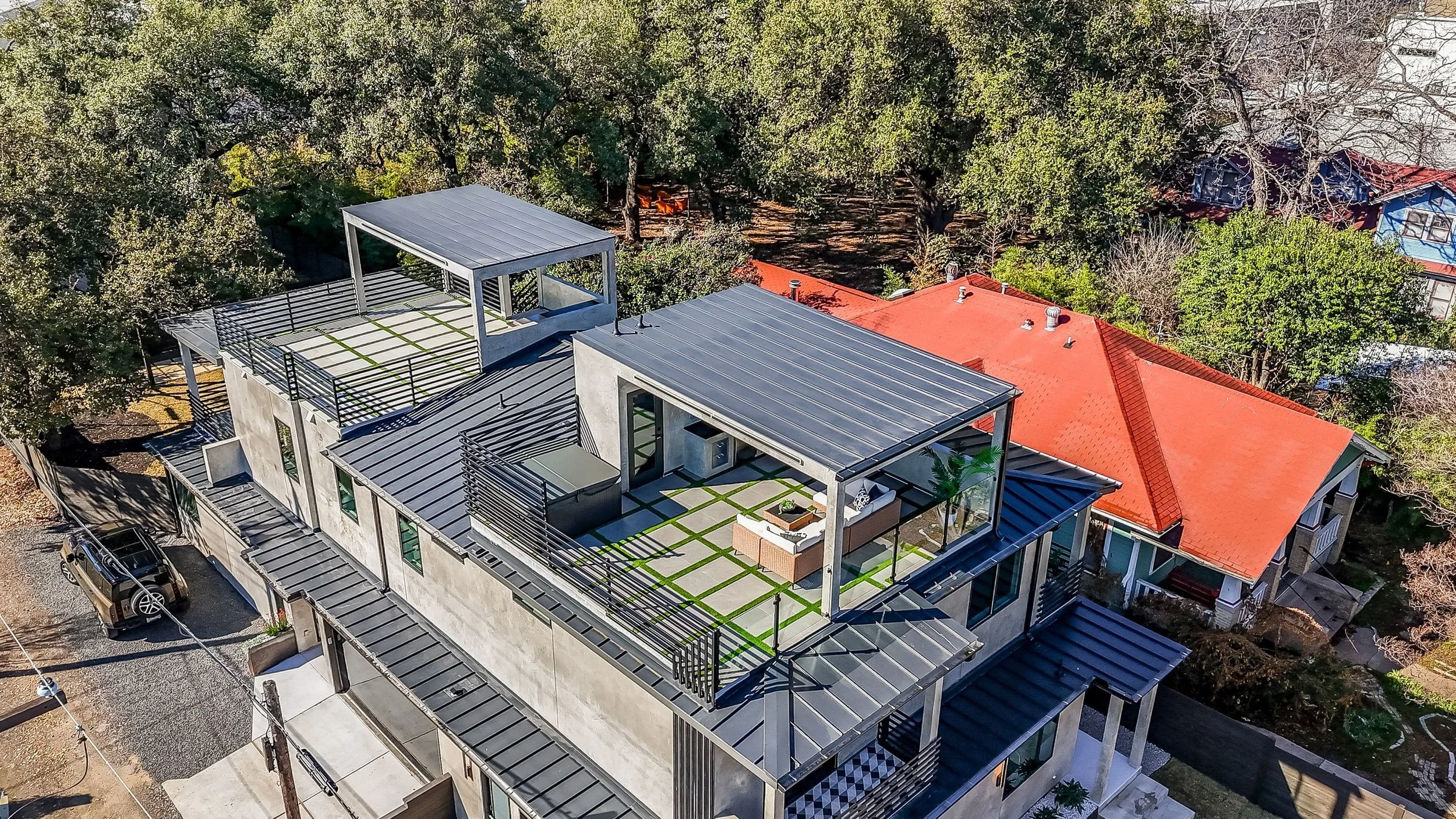Aerial view of a modern house with rooftop patio, outdoor furniture, and green landscaping surrounded by trees and neighboring houses with red and blue roofs.