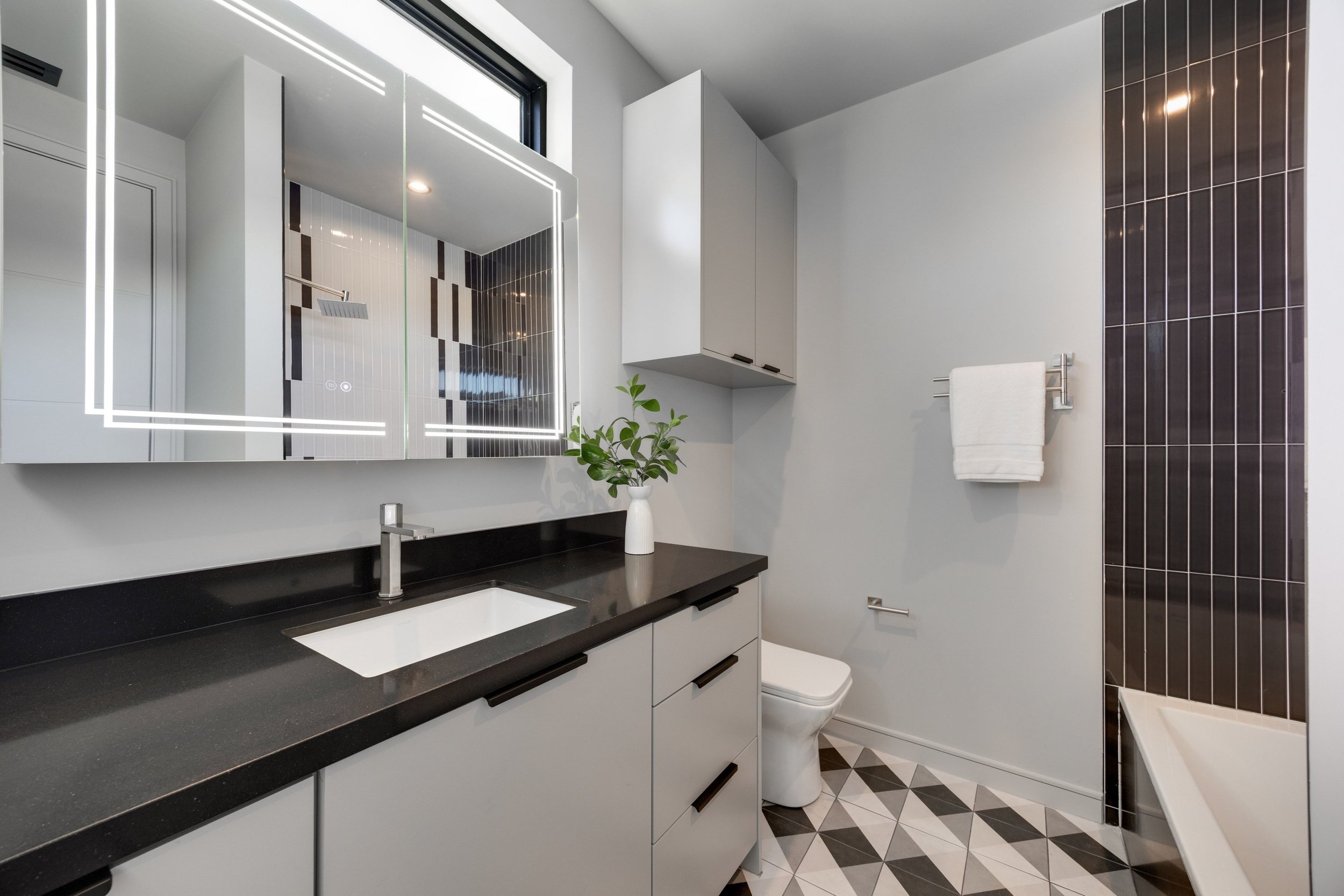 Modern bathroom with a black countertop, white cabinetry, a mirror with built-in LED lighting, a small faucet, a white vase with green leaves, a toilet, white walls, a wall-mounted cabinet, a towel rack with a white towel, dark vertical tiles on the 