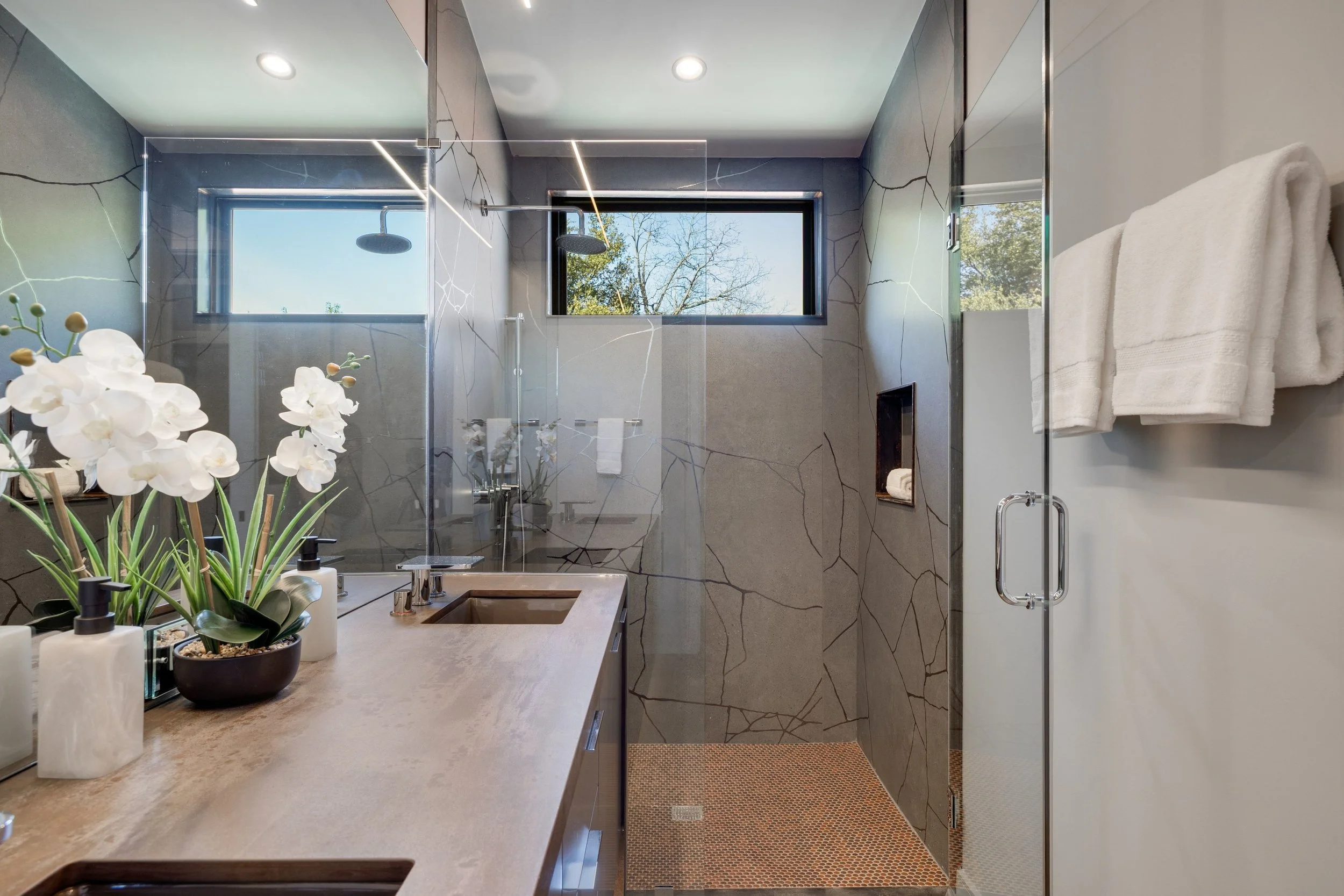 Modern bathroom with a double vanity, white orchids, and a walk-in shower with gray marble walls and two windows showing trees.