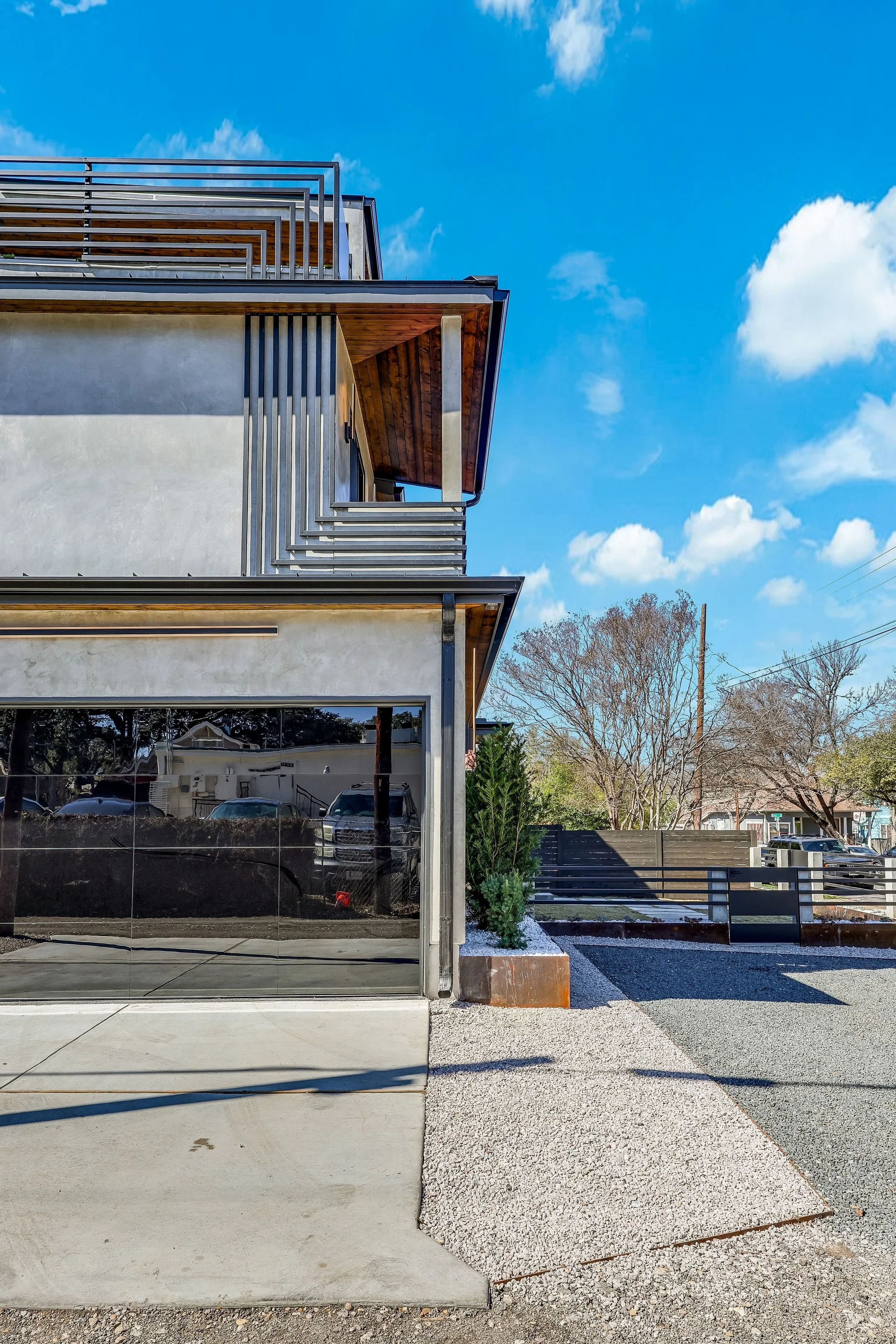 Modern building with glass garage door, metal accents, wooden overhead sections, and a small landscaped area with a plant, under a partly cloudy blue sky.