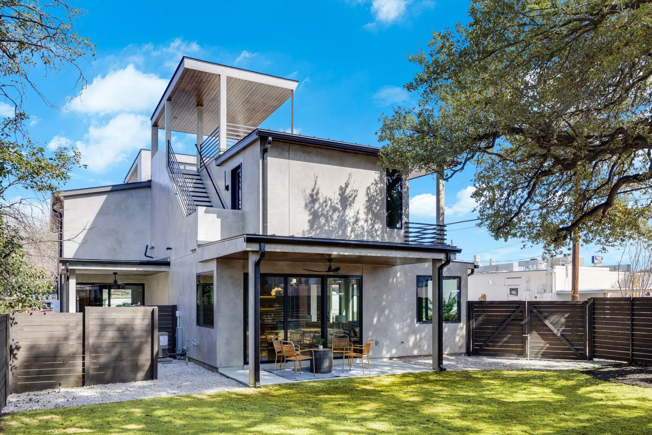 Modern two-story house with a backyard, patio, and outdoor seating area, surrounded by a wooden fence and large trees, under a blue sky with scattered clouds.