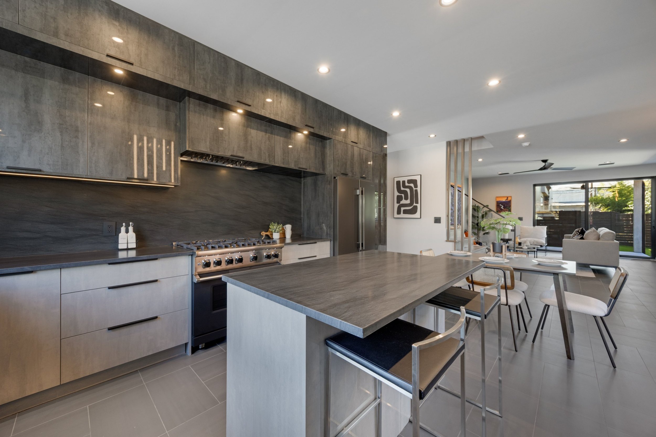Modern kitchen with dark wood cabinets, stainless steel appliances including a large refrigerator and stove, a central island with a wood countertop, and an open living area with white furniture and large windows.
