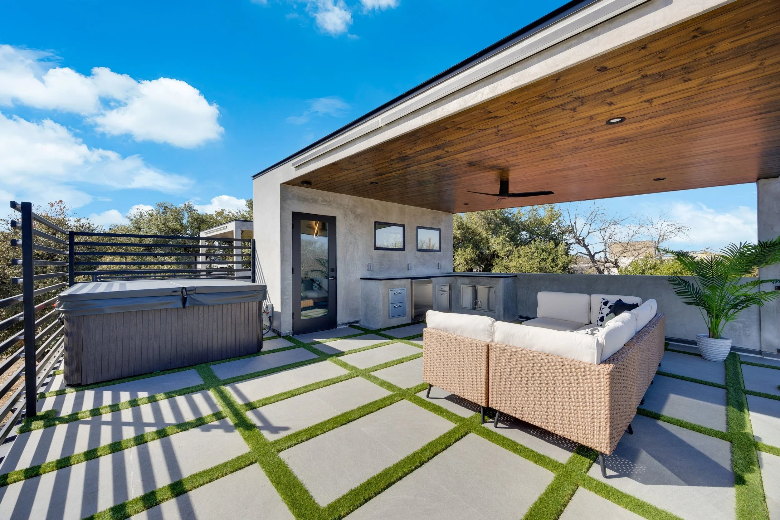 Modern rooftop terrace with outdoor seating, hot tub, built-in bar, and potted plant under a wooden ceiling and blue sky.