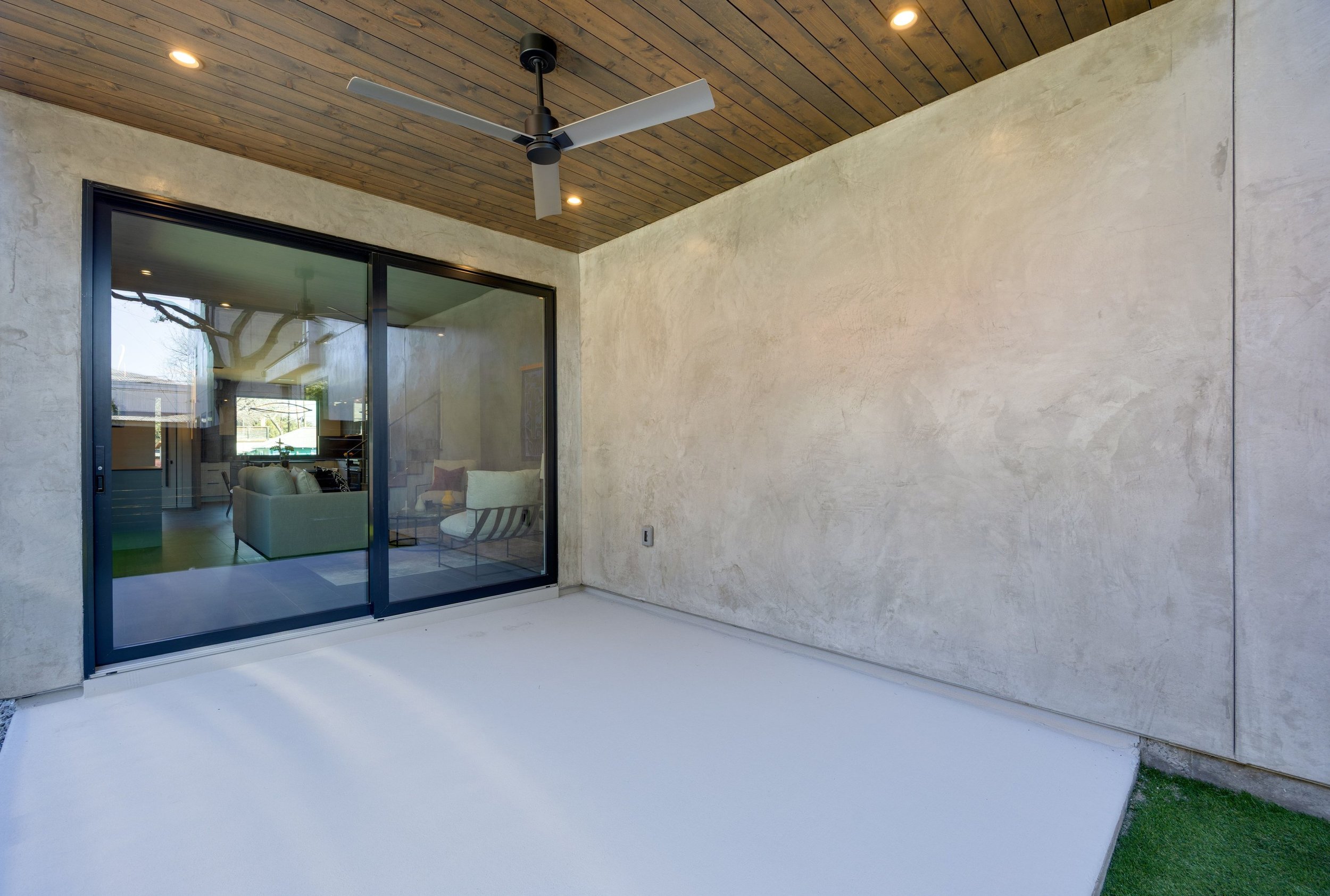 Modern outdoor patio with concrete floor, large glass sliding doors, beige textured walls, and a wooden ceiling with recessed lighting and a ceiling fan.