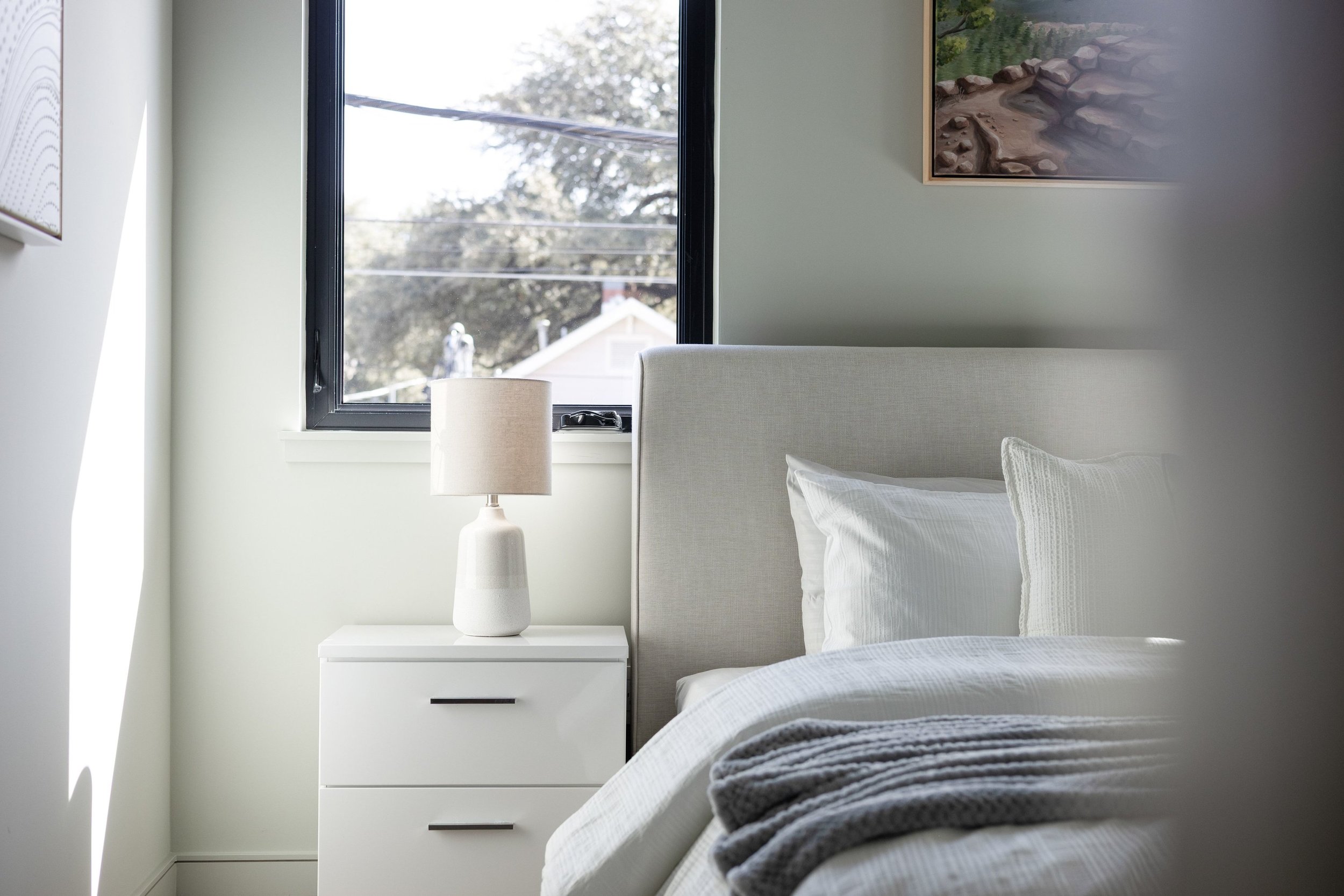 A neatly made bed with white linens and pillows, a white nightstand with a white table lamp, a window showing a sunny outdoor scene with trees, and a painting on the wall of a landscape.