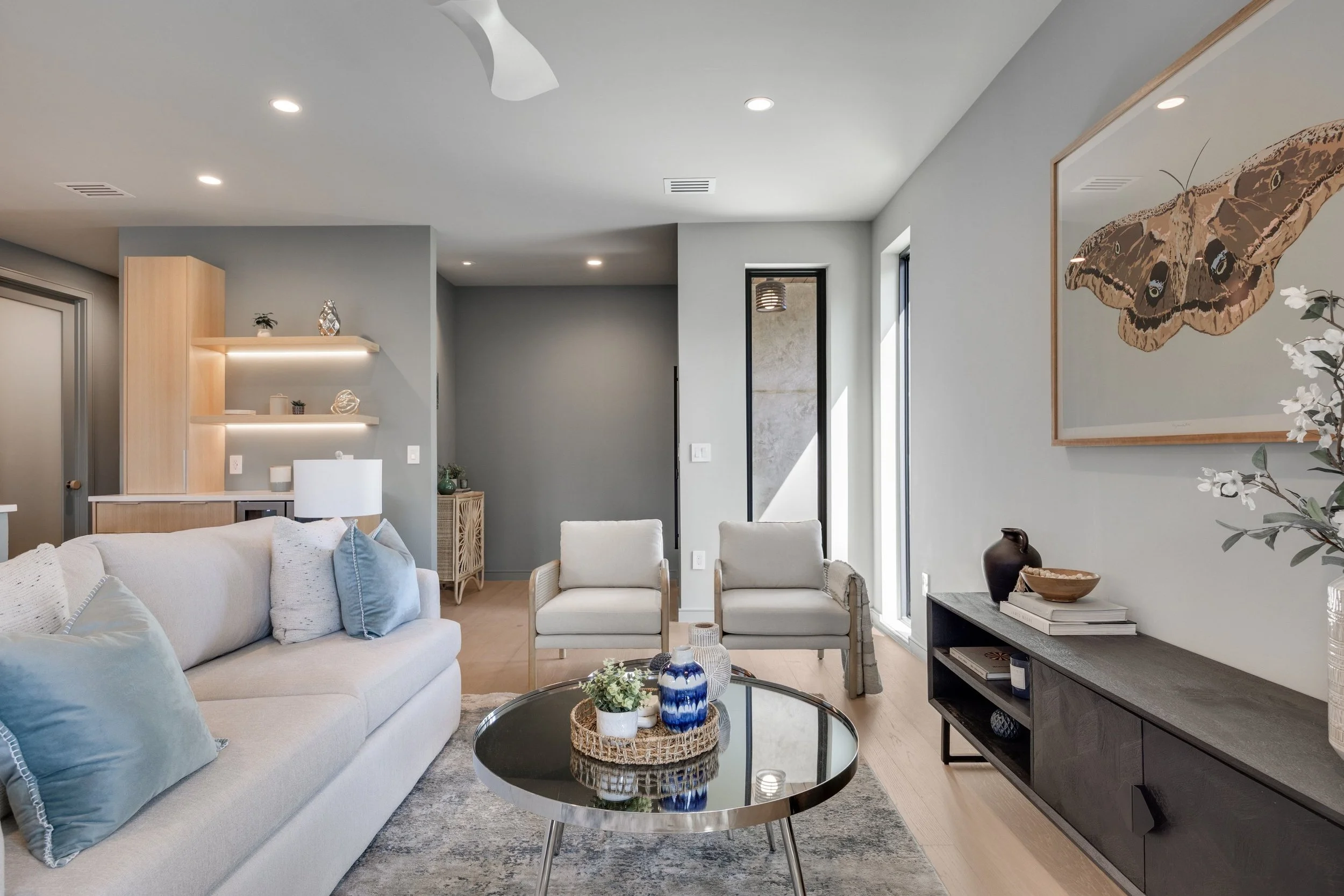 A modern living room with a white sofa and two light-colored chairs. A round glass coffee table with decorative items is in the center. A long black TV stand holds decor, and a framed butterfly artwork hangs on the light gray wall. There's a window l