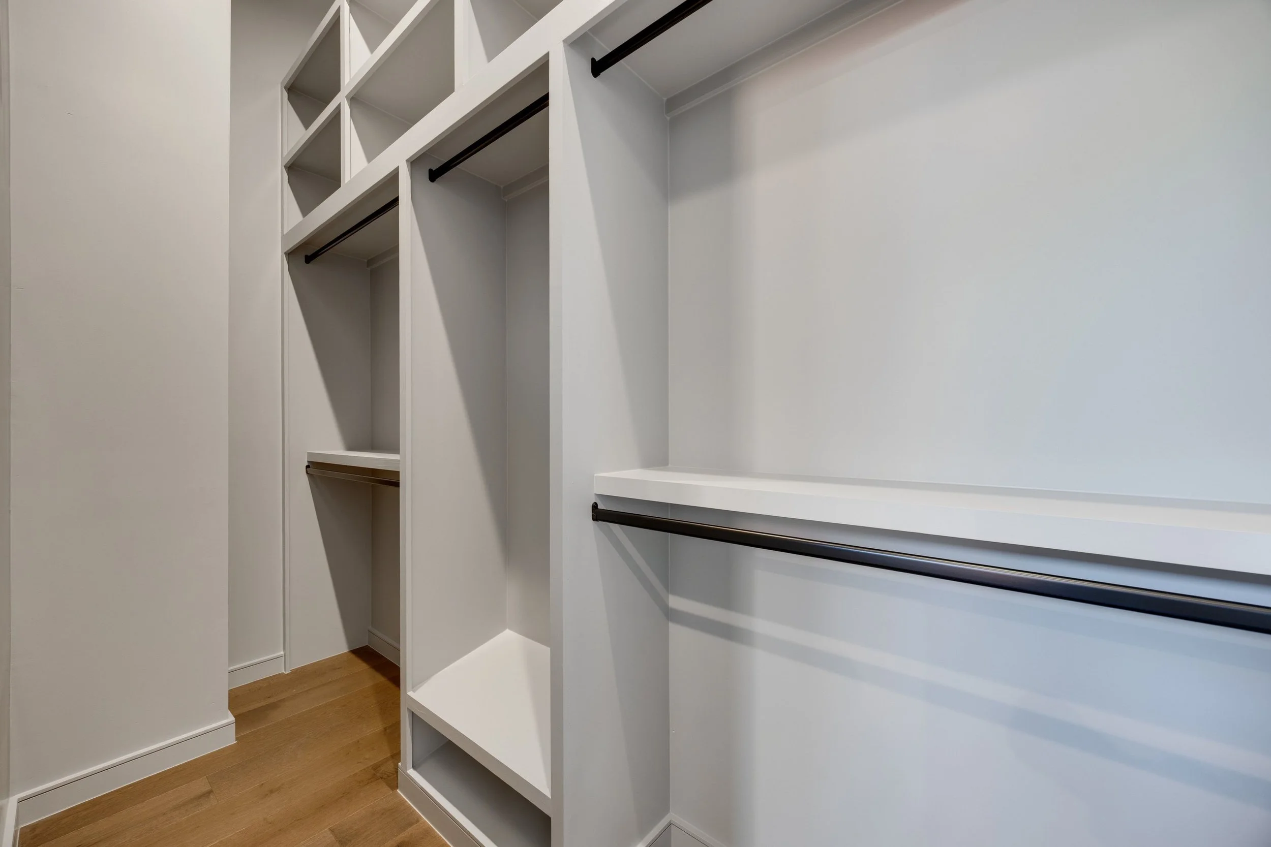 Empty walk-in closet with white built-in shelves and black hanging rods, hardwood floor.