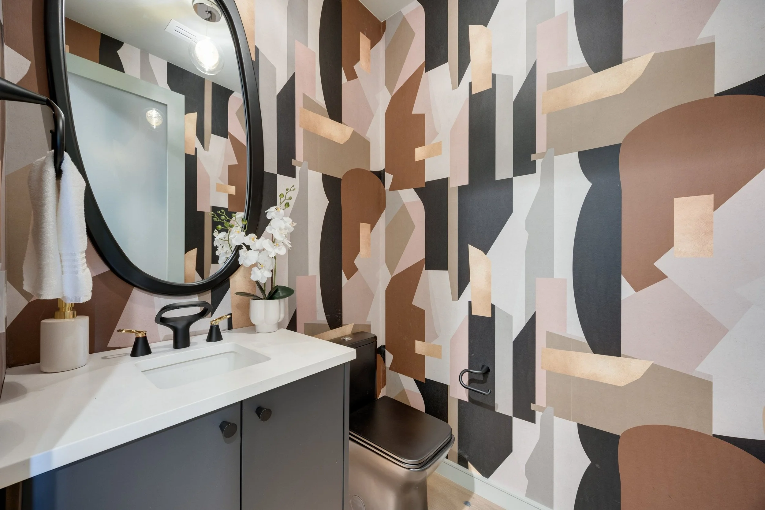 Modern bathroom with abstract geometric wall mural, oval mirror, black sink faucet, white countertop, vase with white orchids, small hand towel on a dispenser, and black toilet.
