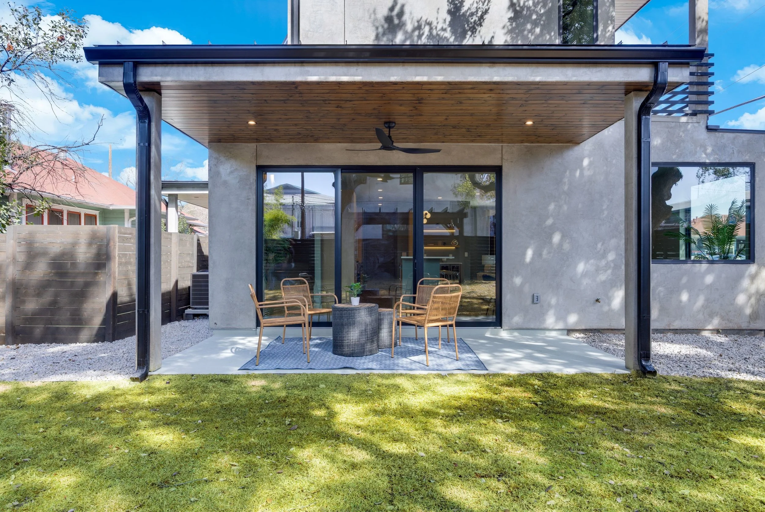 The image shows a modern outdoor patio with four chairs around a small round table on a rug, in front of a house with large sliding glass doors and windows, under a wooden overhang with a ceiling fan, on a grassy yard with a fence and neighboring hou