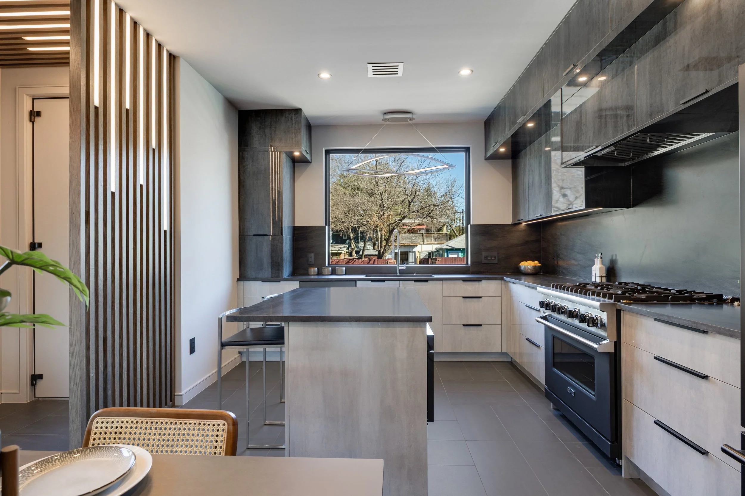 Modern kitchen with gray cabinets, a large window, a black stove, a kitchen island with a gray countertop, and a dining table with a plate and a woven chair.