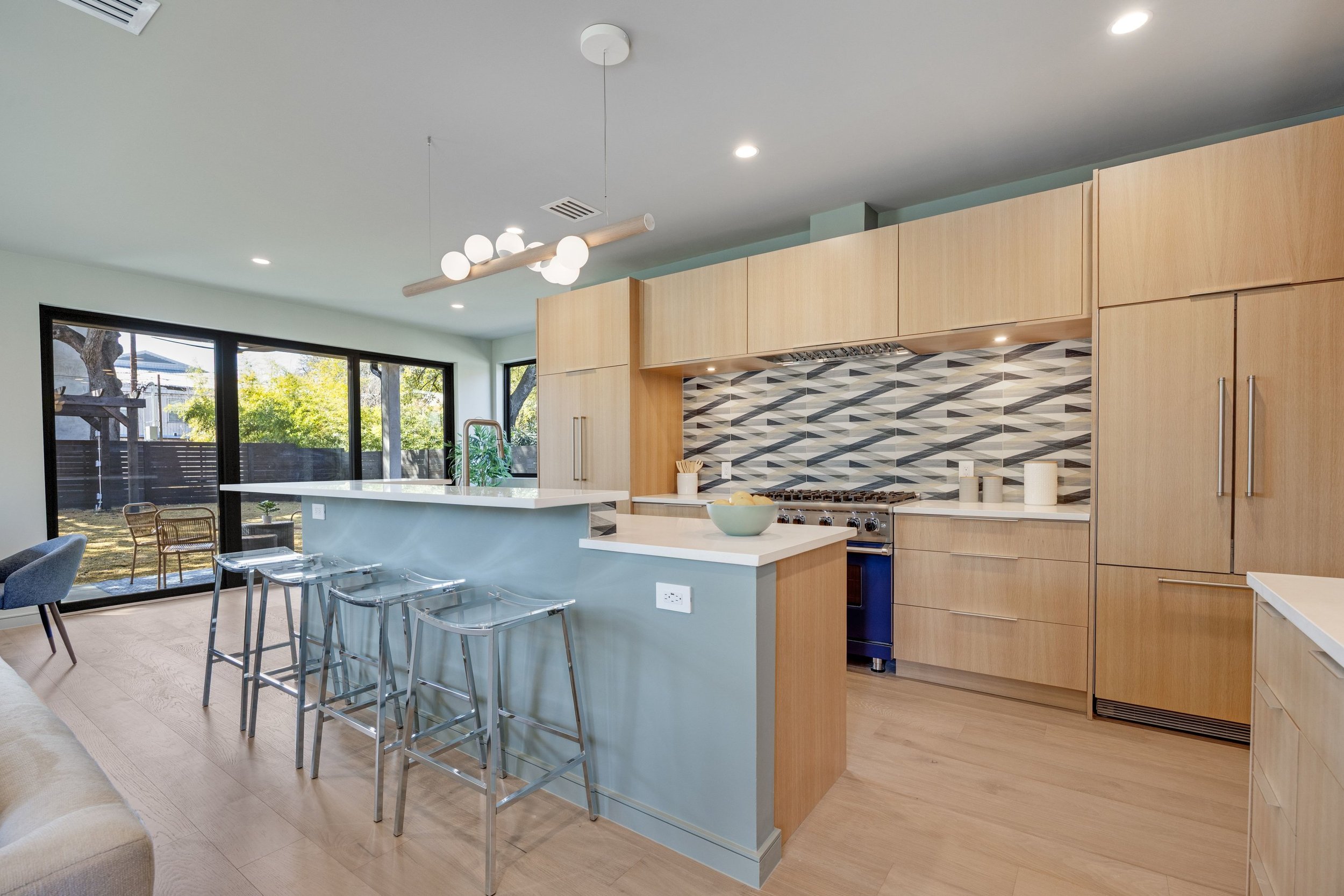 Modern kitchen with light wood cabinets, patterned gray and white backsplash, blue island with seating, large sliding glass doors showing outdoor patio with chairs and trees.
