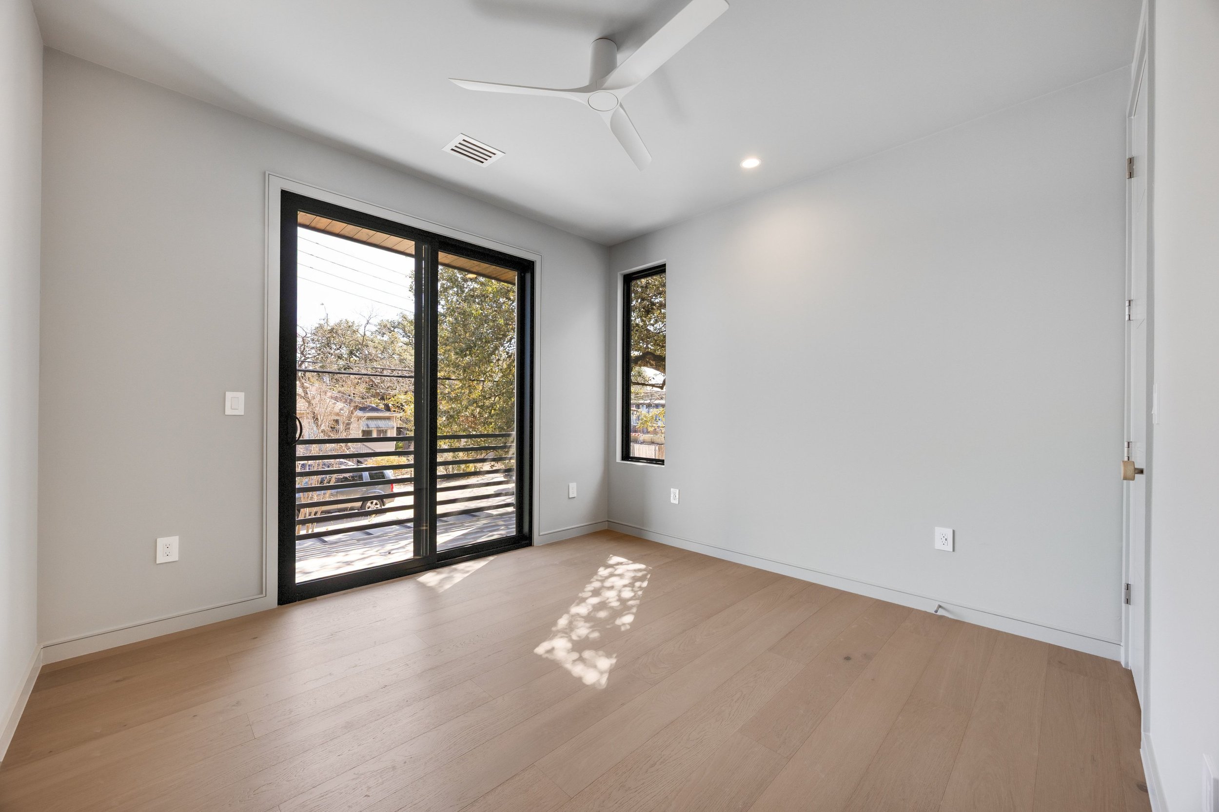 Empty room with wooden floors, white walls, a sliding glass door leading to a balcony, and a ceiling fan.