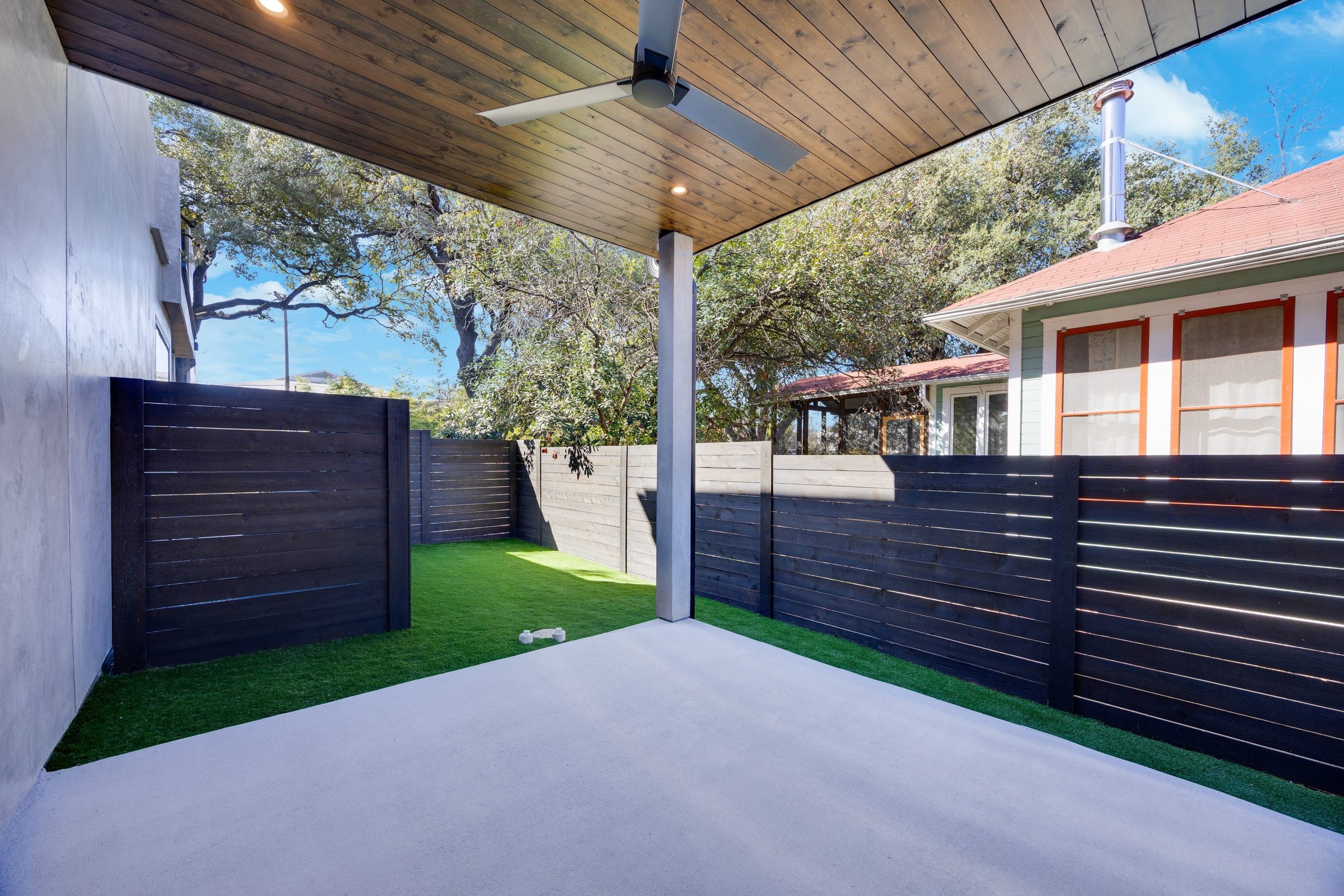 View of a backyard patio with a concrete floor, grassy areas, and black wooden fencing. There is a ceiling fan and wooden ceiling on the porch area, with trees and neighboring houses in the background.