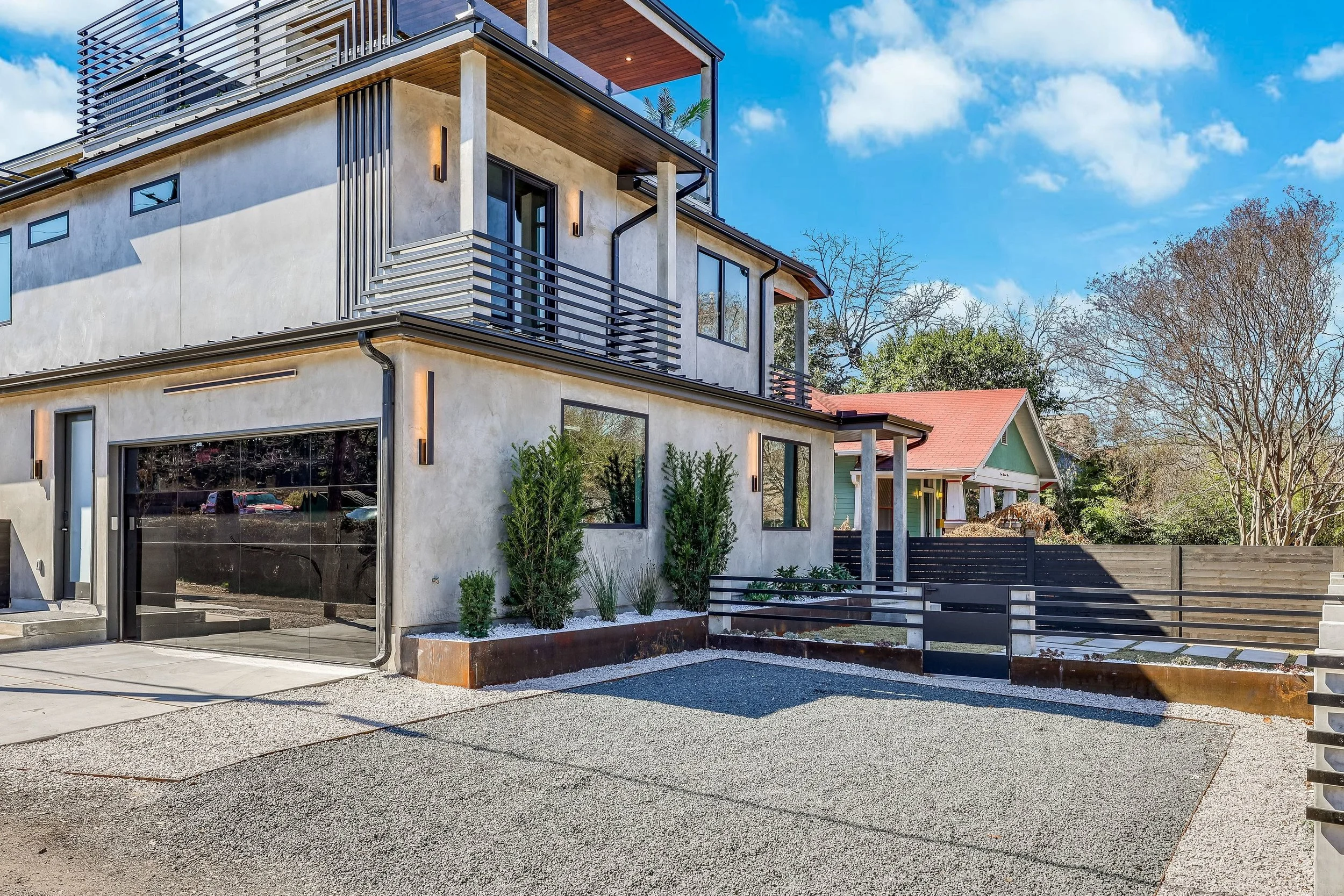 Modern multi-story house with concrete and wood exterior, large glass windows, metal railings, and a small landscaped yard with bushes. Blue sky with clouds in the background.