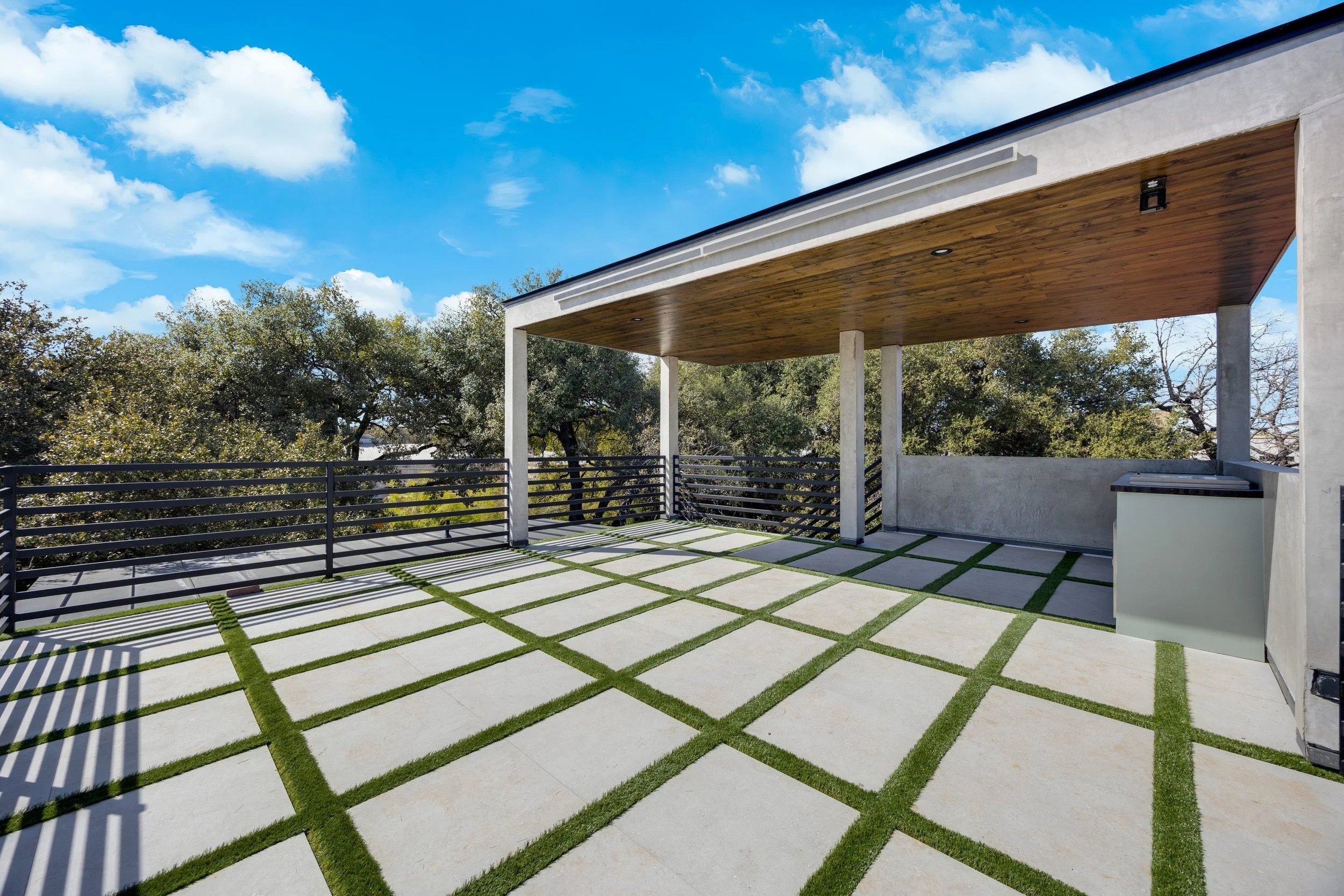Modern outdoor rooftop patio with concrete pavers and grass strips, black metal railing, wooden ceiling, and scenic view of trees under a blue sky with clouds.