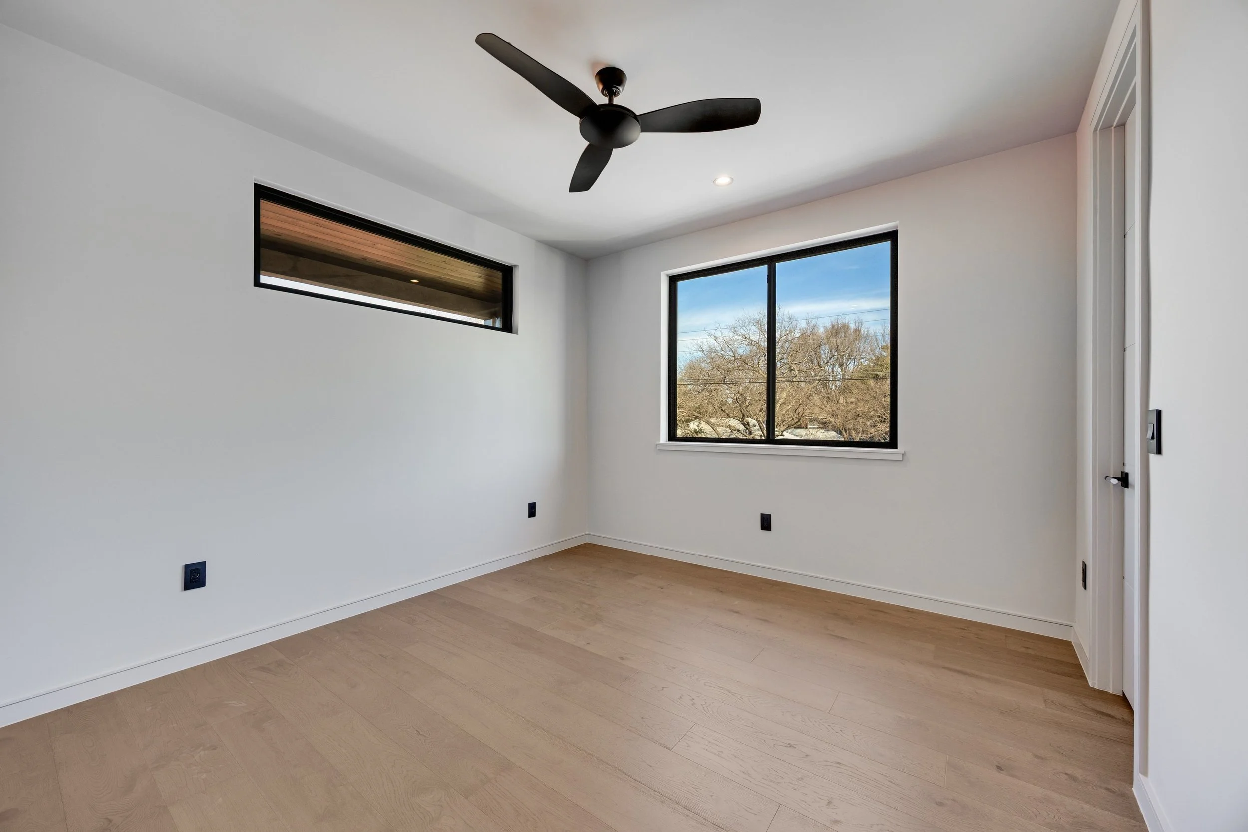 Empty room with white walls, light wood floor, black ceiling fan, two black-framed windows showing trees outside, and a door on the right.