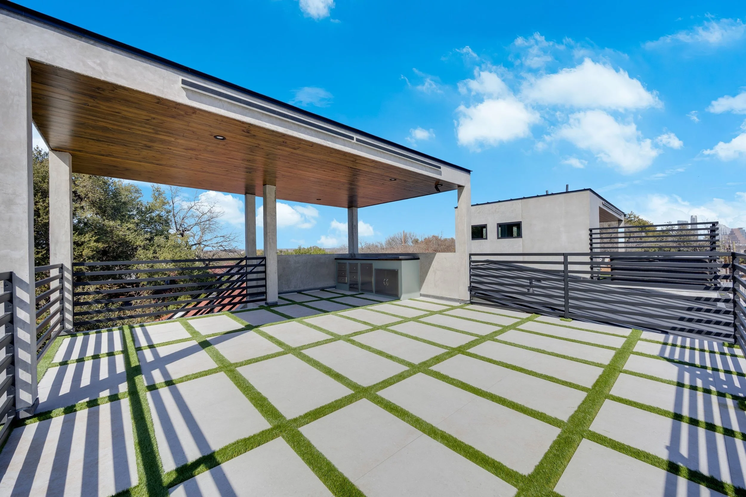 Modern rooftop terrace with concrete flooring, grass strips, black metal railings, a covered area with wooden ceiling, and cityscape in the background under a blue sky with clouds.