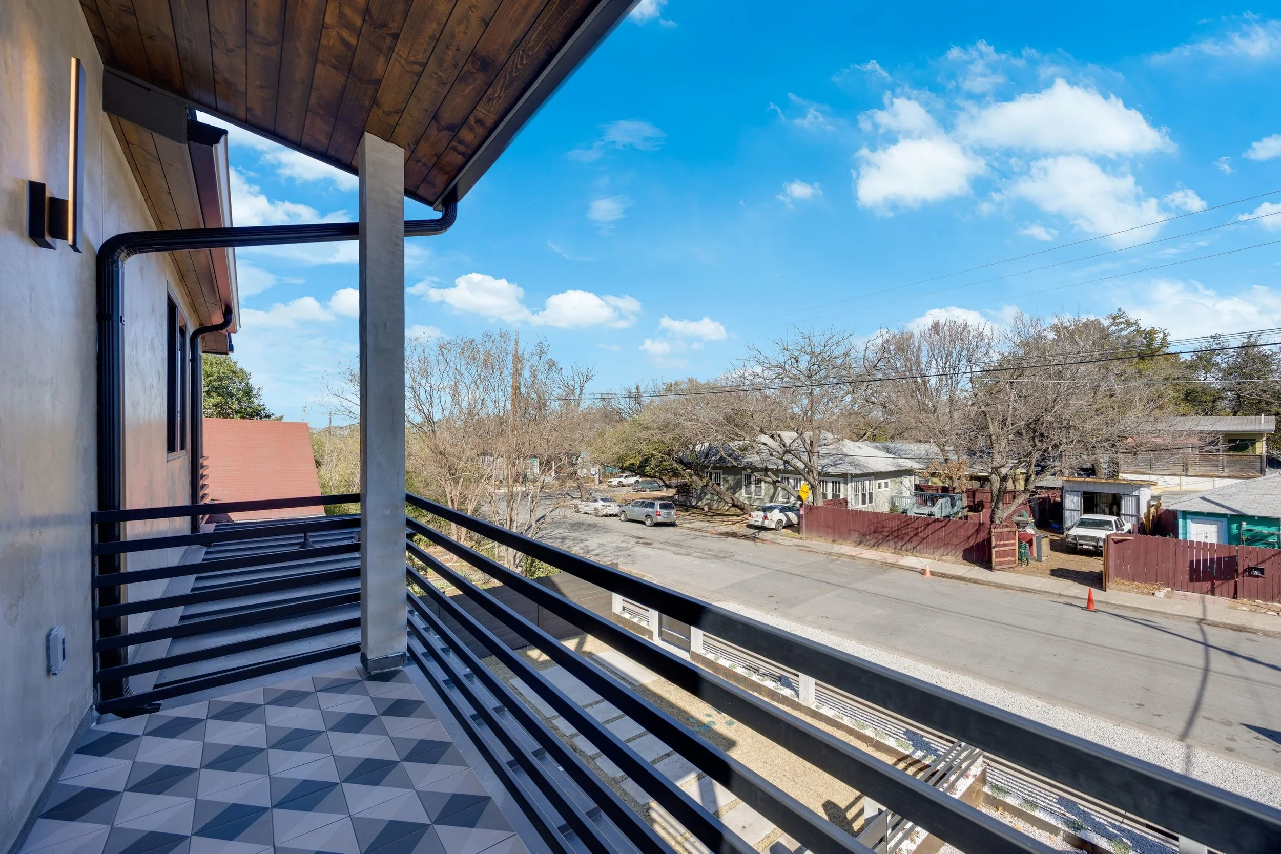 View from a balcony overlooking a neighborhood street with parked cars, leafless trees, residential houses, and a bright blue sky with scattered white clouds.