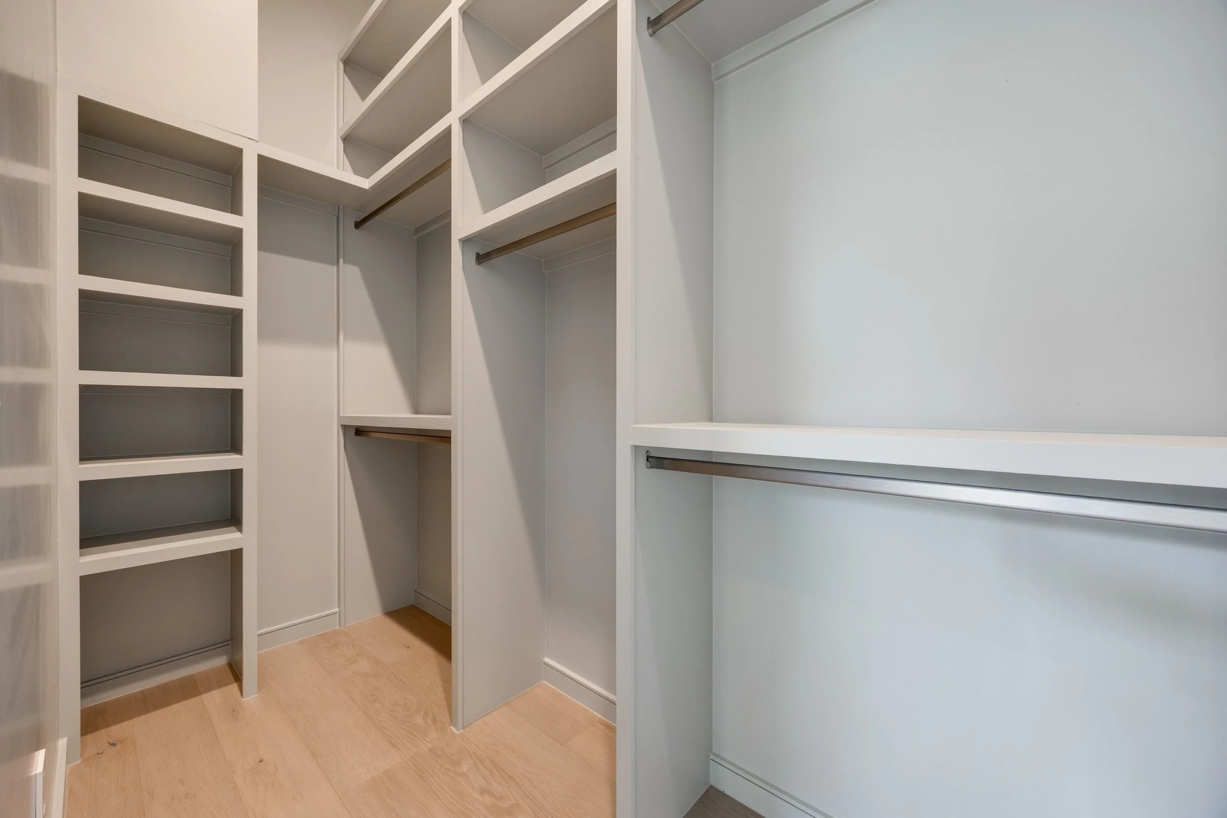 Empty walk-in closet with white shelving, hanging rods, and light wood floor.