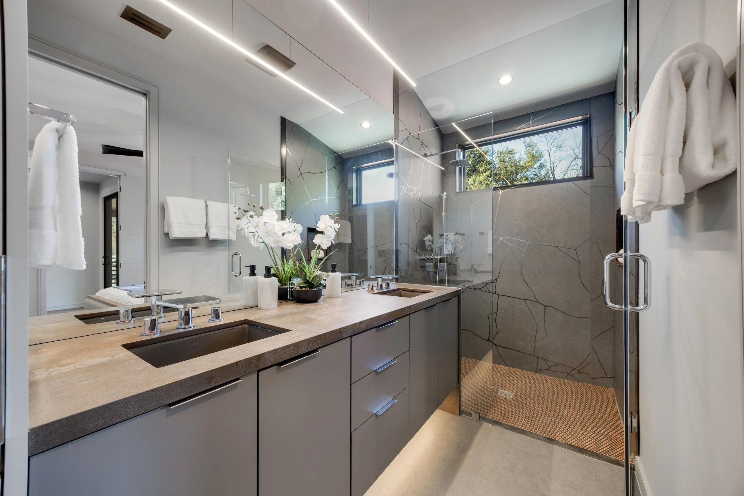 Modern bathroom with double vanity, large mirror, flower arrangement, and a walk-in shower with glass door and window.