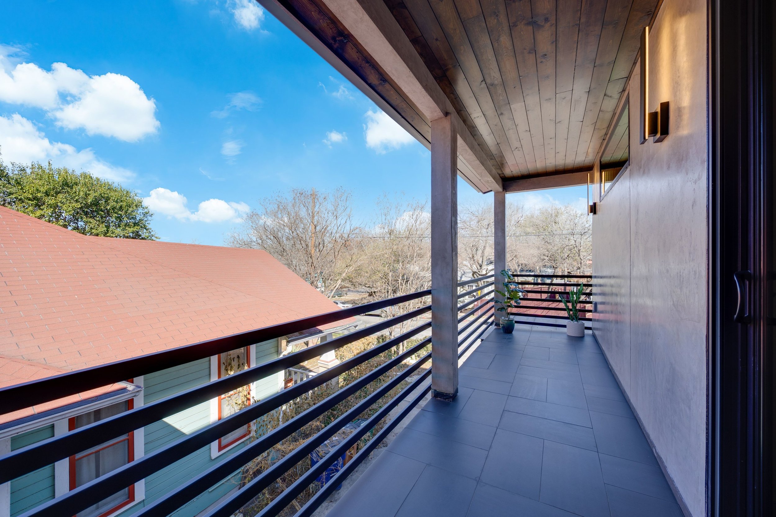 Balcony with gray tiled floor, potted plants, wooden ceiling, and black metal railing, overlooking a neighboring house with a red shingle roof and trees under a blue sky with clouds.