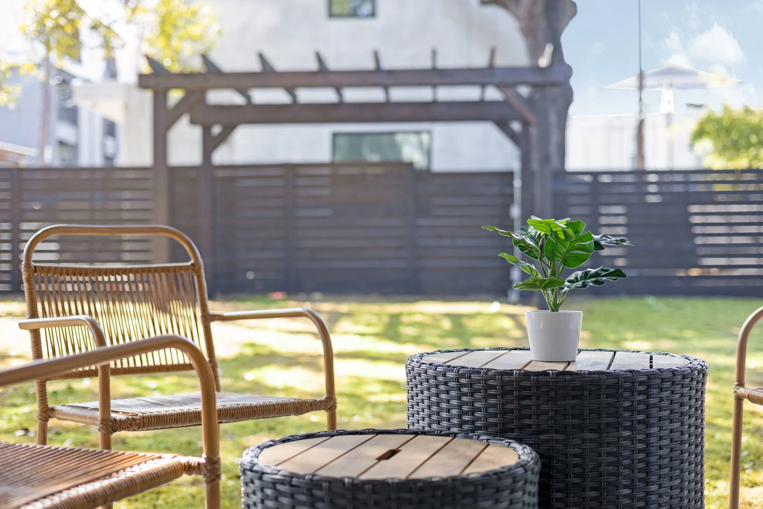 A backyard patio area with wicker chairs, a wicker table with a potted plant, and a wooden fence in the background. There's an umbrella and trees visible in the distance.
