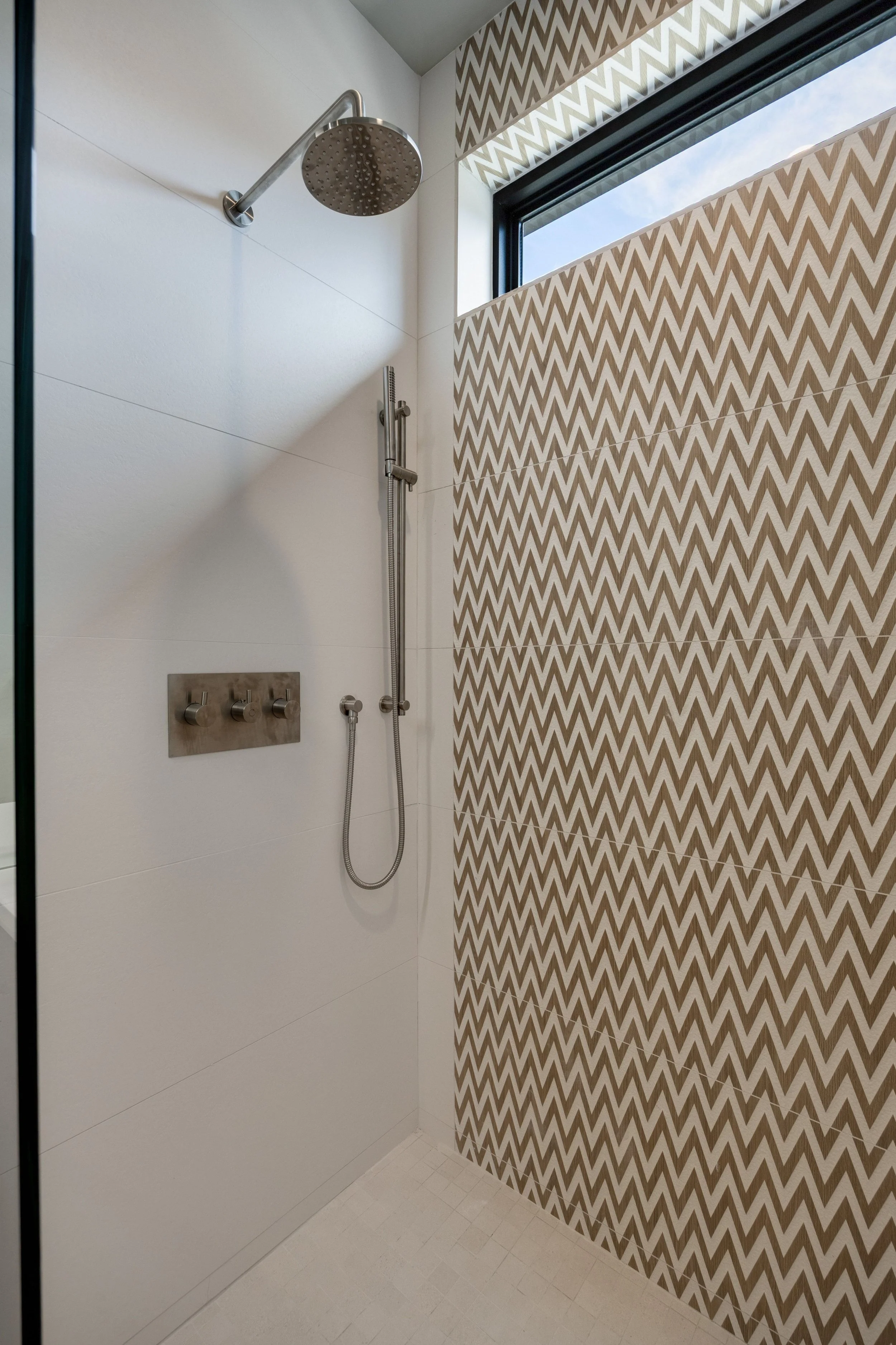 A modern shower with a large rain showerhead, a hand-held shower, and a textured wall with brown and white chevron pattern near a high window.