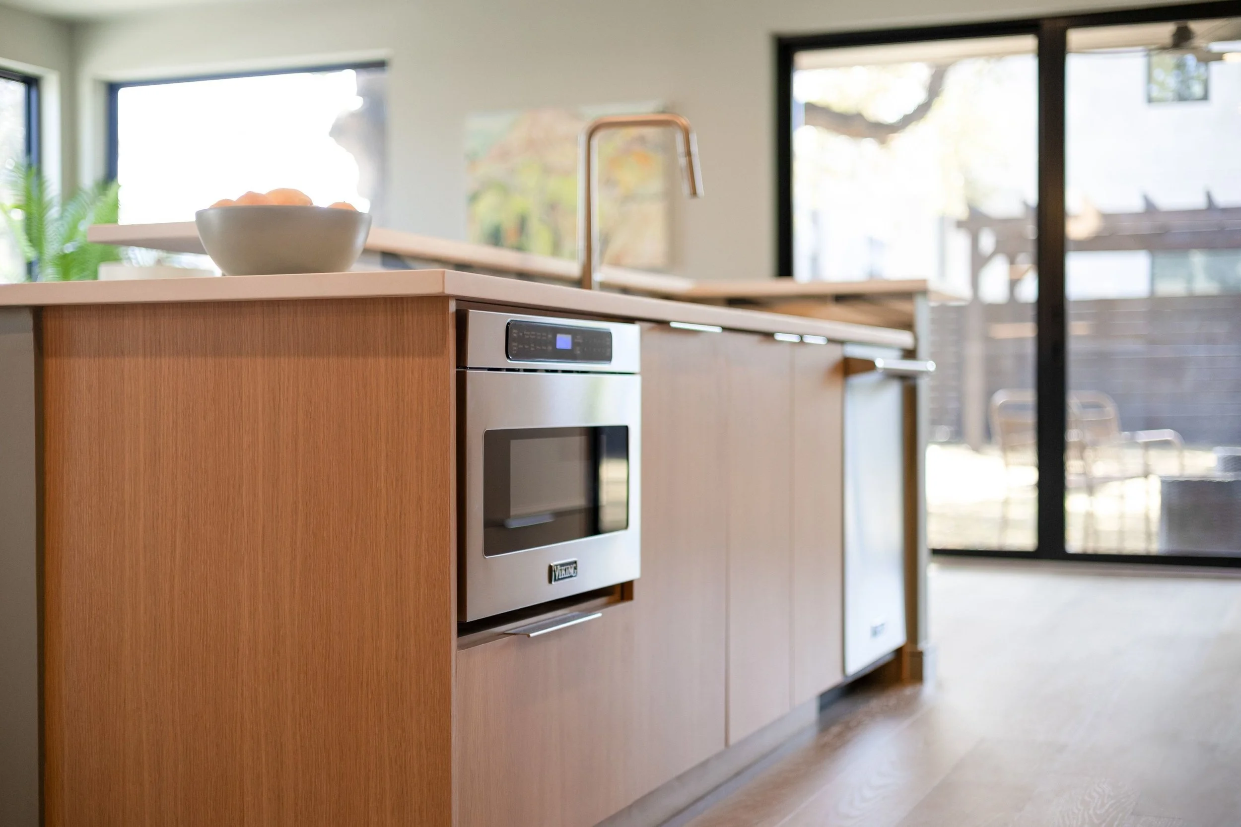 Modern kitchen with light wood cabinetry, a built-in microwave, a white countertop, a bowl of fruit, and large glass sliding doors leading outside.