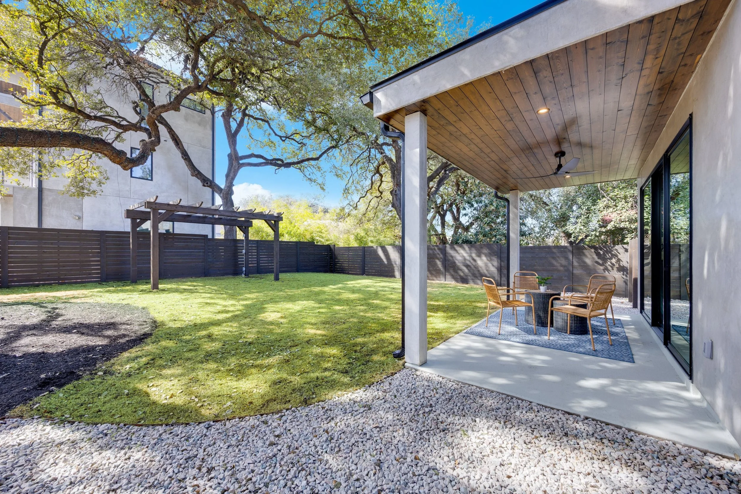 View of a backyard with a covered patio area furnished with a round table and four chairs, a grassy yard surrounded by a tall black fence, a large tree with spreading branches, and neighboring buildings in the background. The sky is clear and sunny.