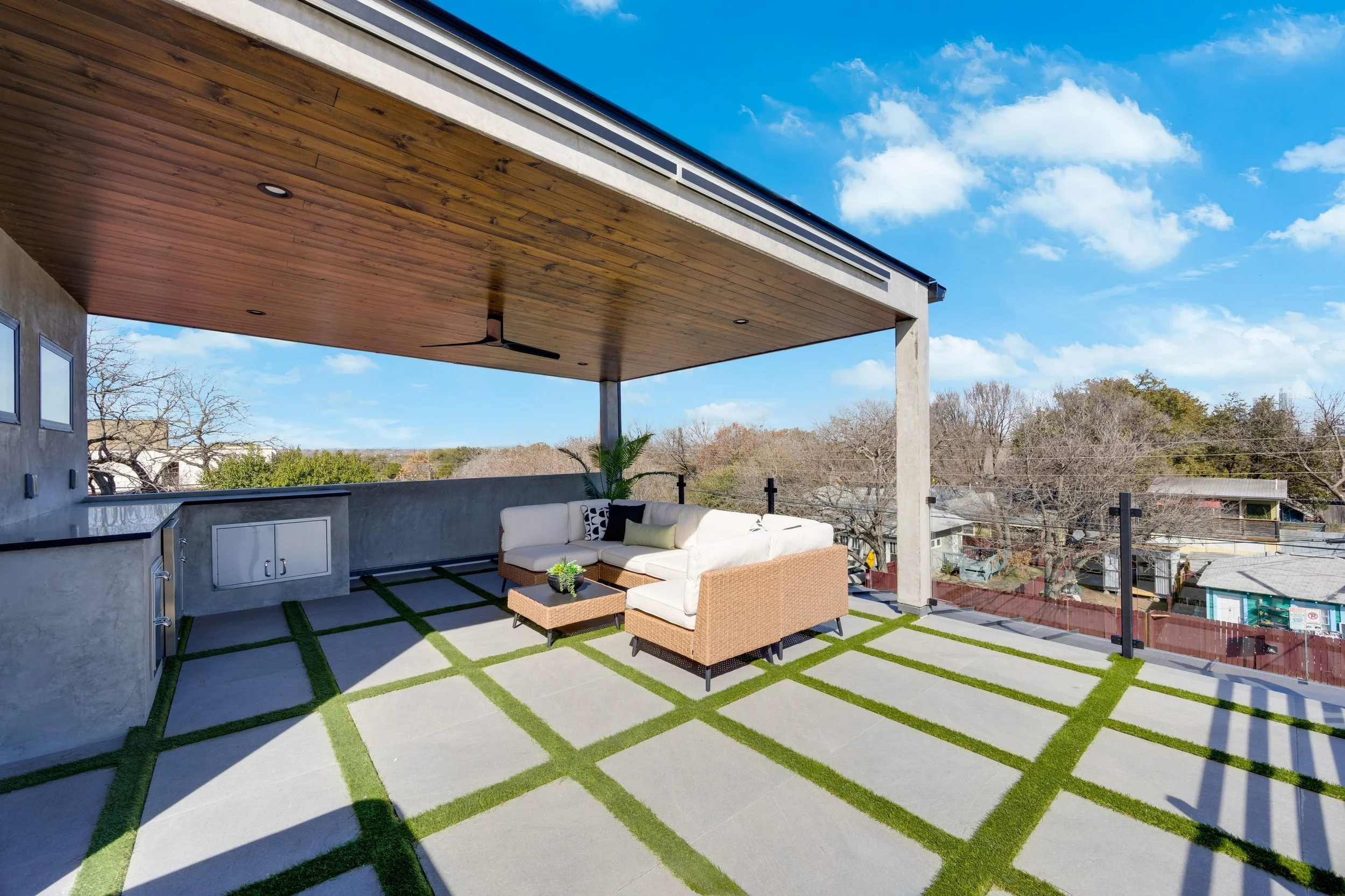 Outdoor rooftop patio with a beige sectional sofa, a small coffee table, and decorative pillows, under a wooden ceiling with a ceiling fan, overlooking a neighborhood with trees and houses under a blue sky with clouds.